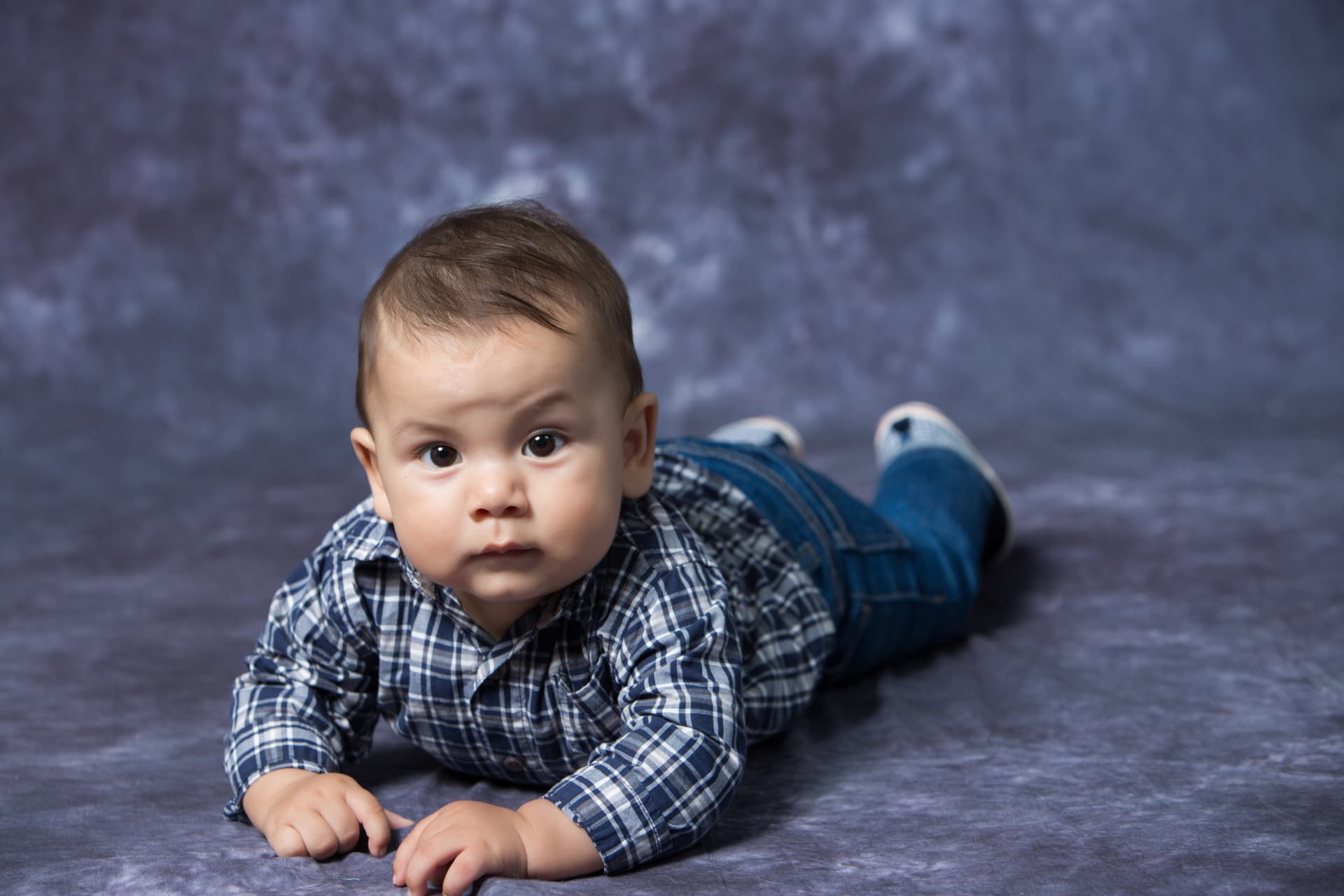 Baby boy in a blue plaid shirt lying against a deep blue studio backdrop