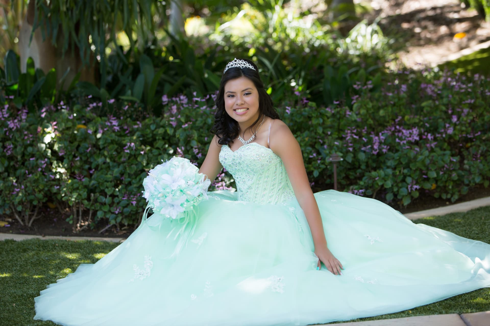 Quinceañera in a mint green gown with tiara and white bouquet seated in a flower garden