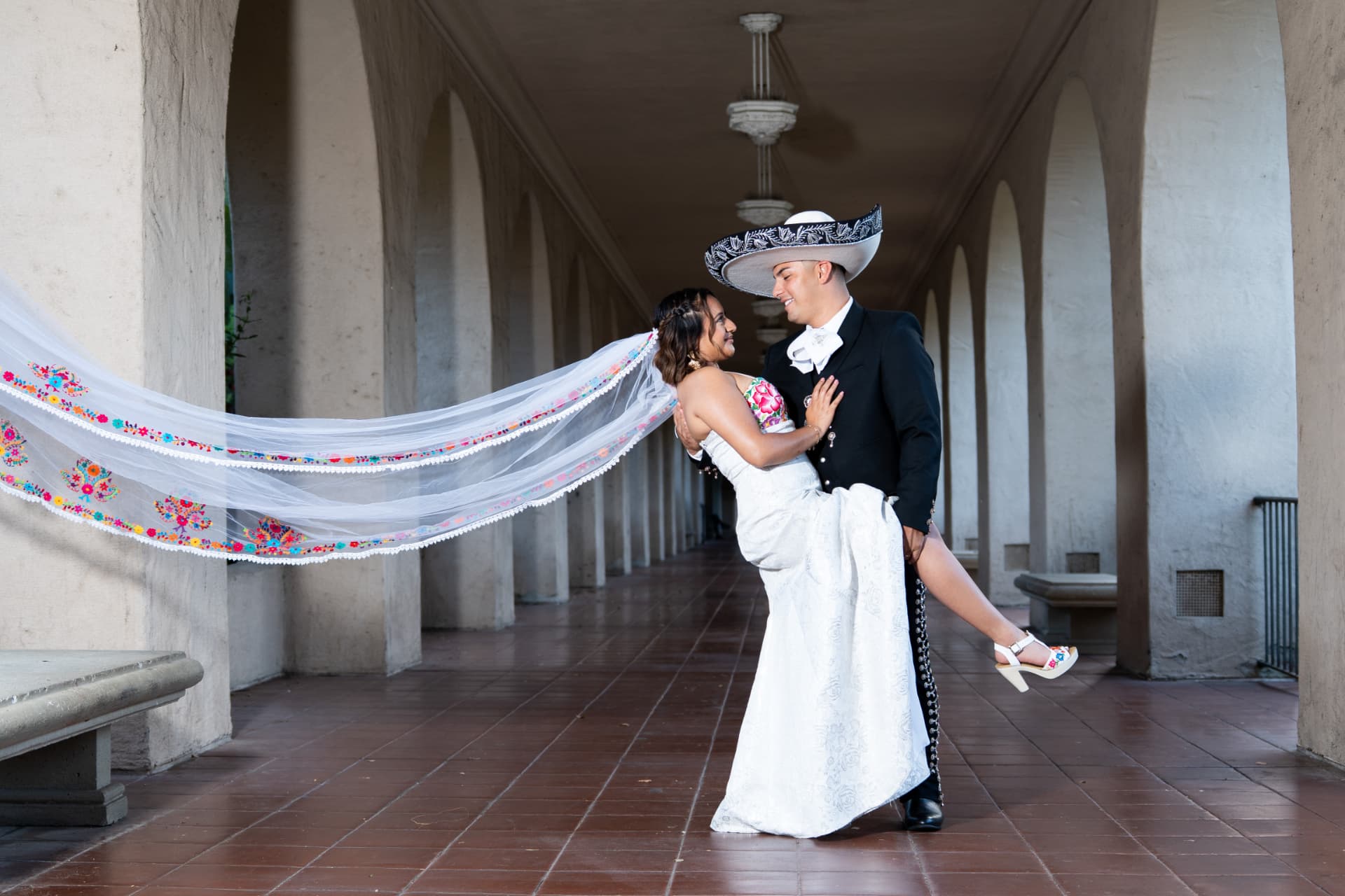 Groom in a sombrero de charro dips his bride as her embroidered veil sweeps through an arched corridor