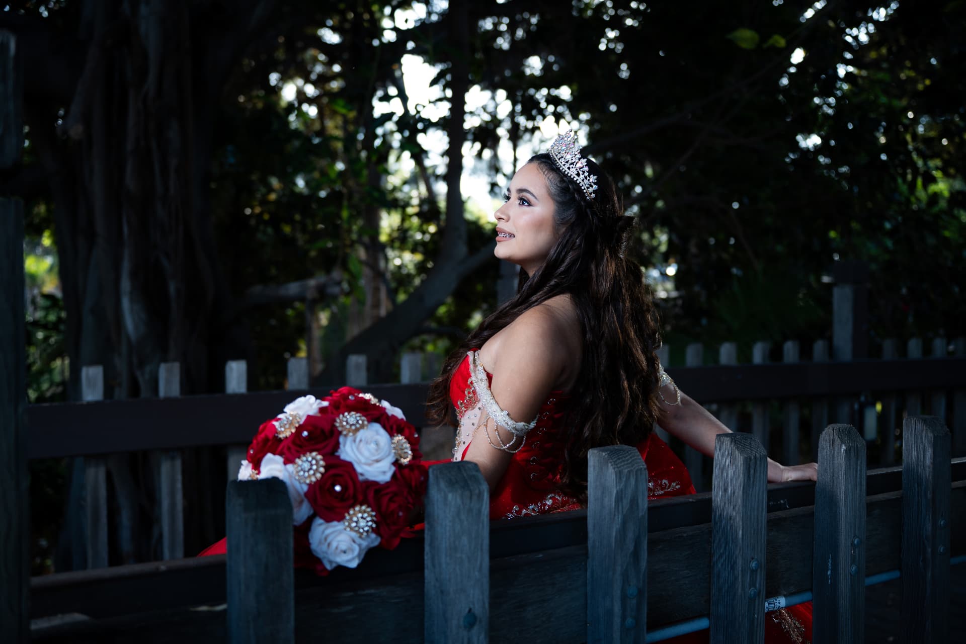Quinceañera in a red gown seated on a wooden bridge with her red and white bouquet