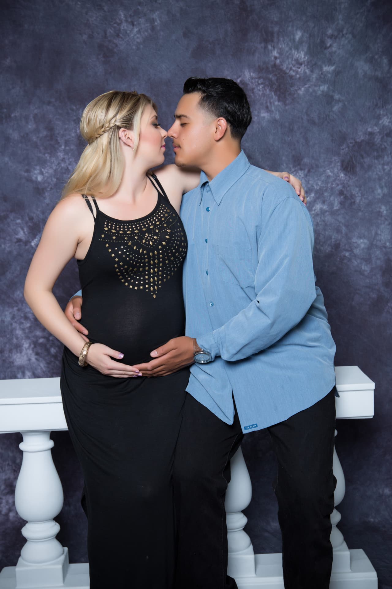 Expectant couple sharing a kiss beside a white studio railing