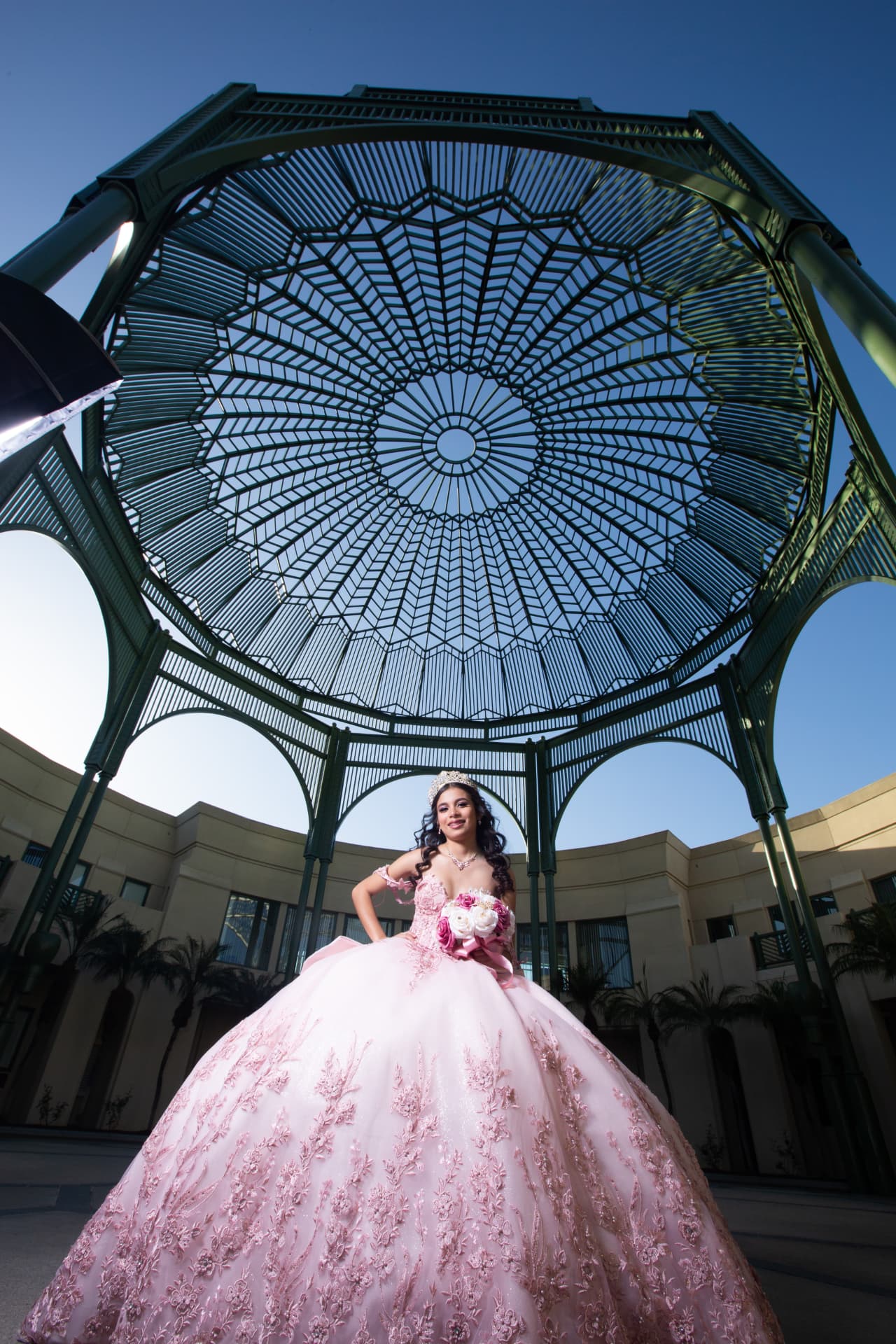 Quinceañera in a blush gown with a bouquet beneath a green lattice gazebo against a blue sky