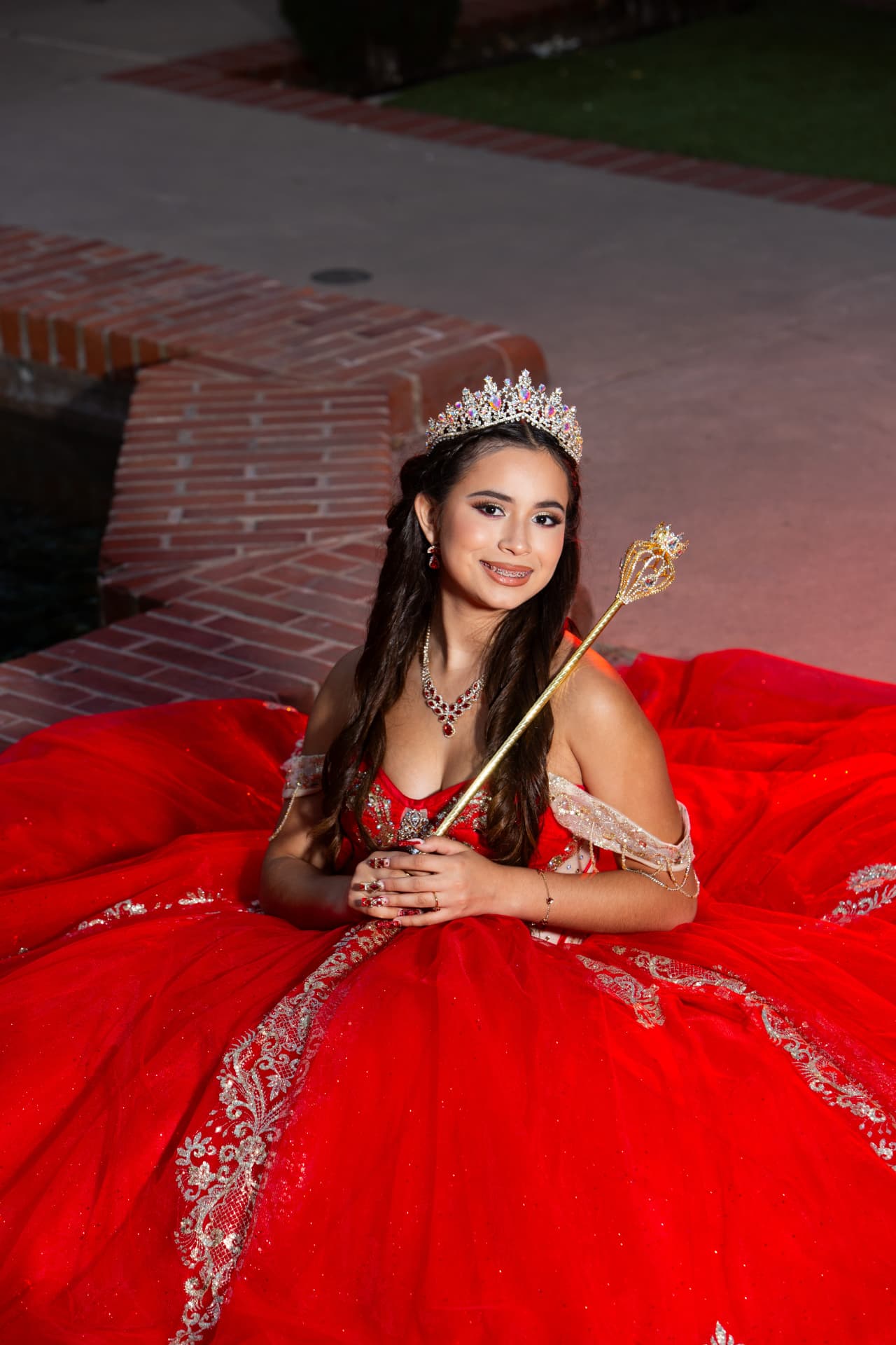 Quinceañera in a red gown holding her golden scepter on a brick pathway