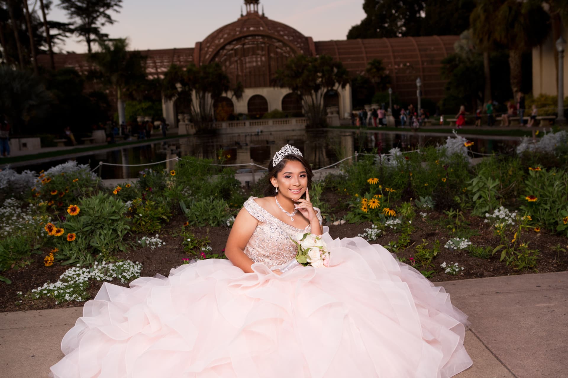 Quinceañera in a pale pink gown seated in a flower garden with a lath house and pond behind her