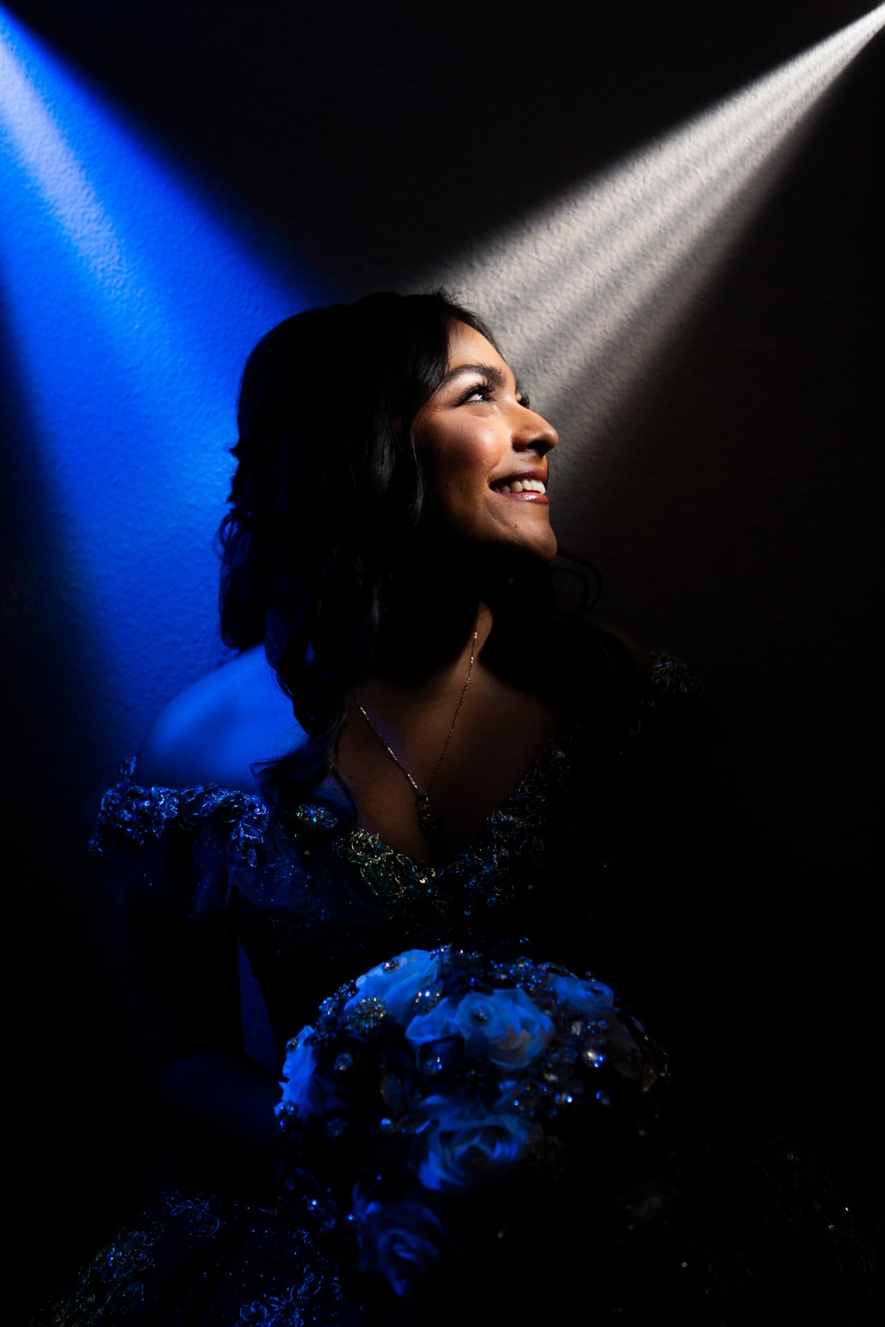 Quinceañera lit by a single blue spotlight, looking up with her bouquet