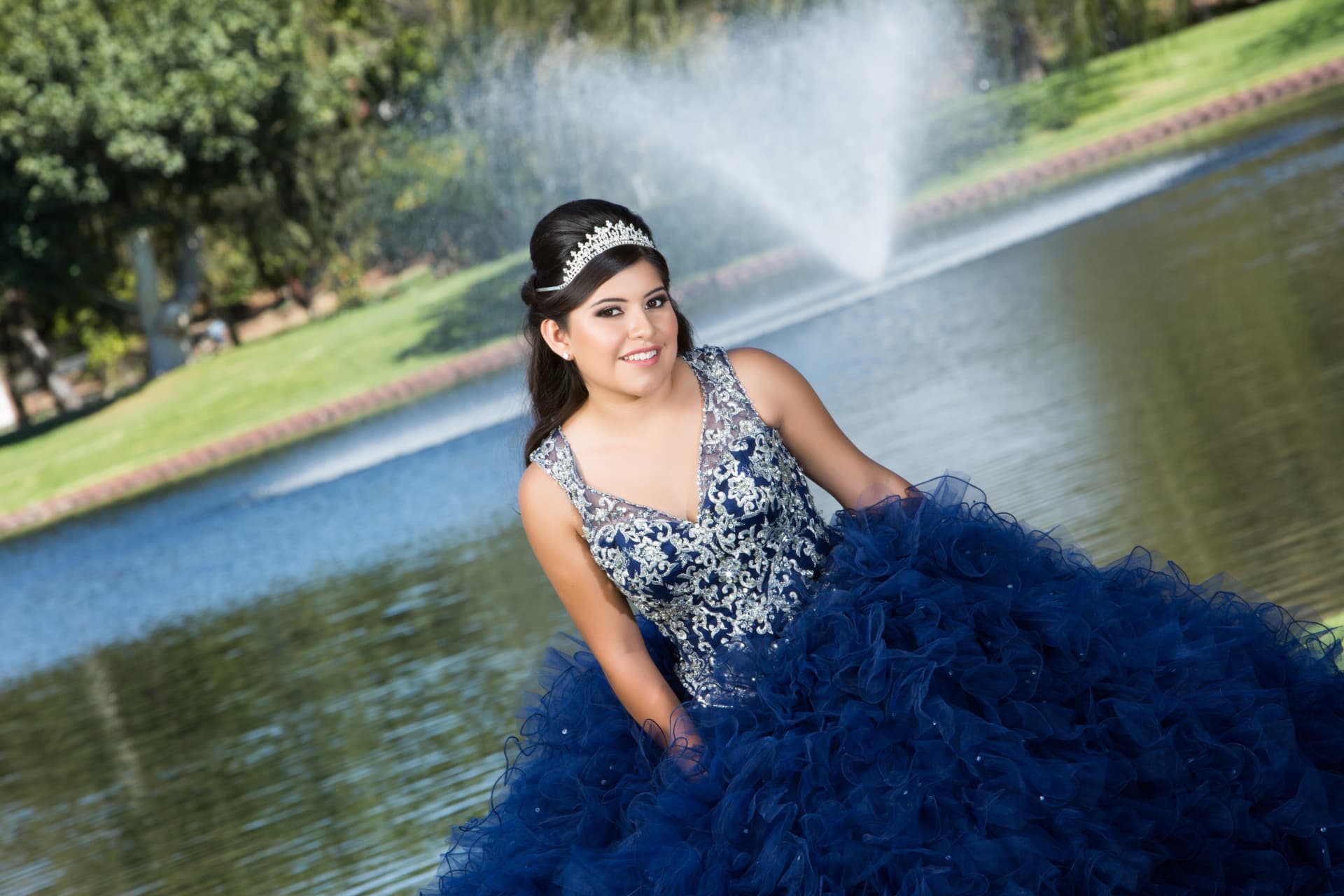 Quinceañera in a navy and silver gown seated by a lake with a fountain behind her