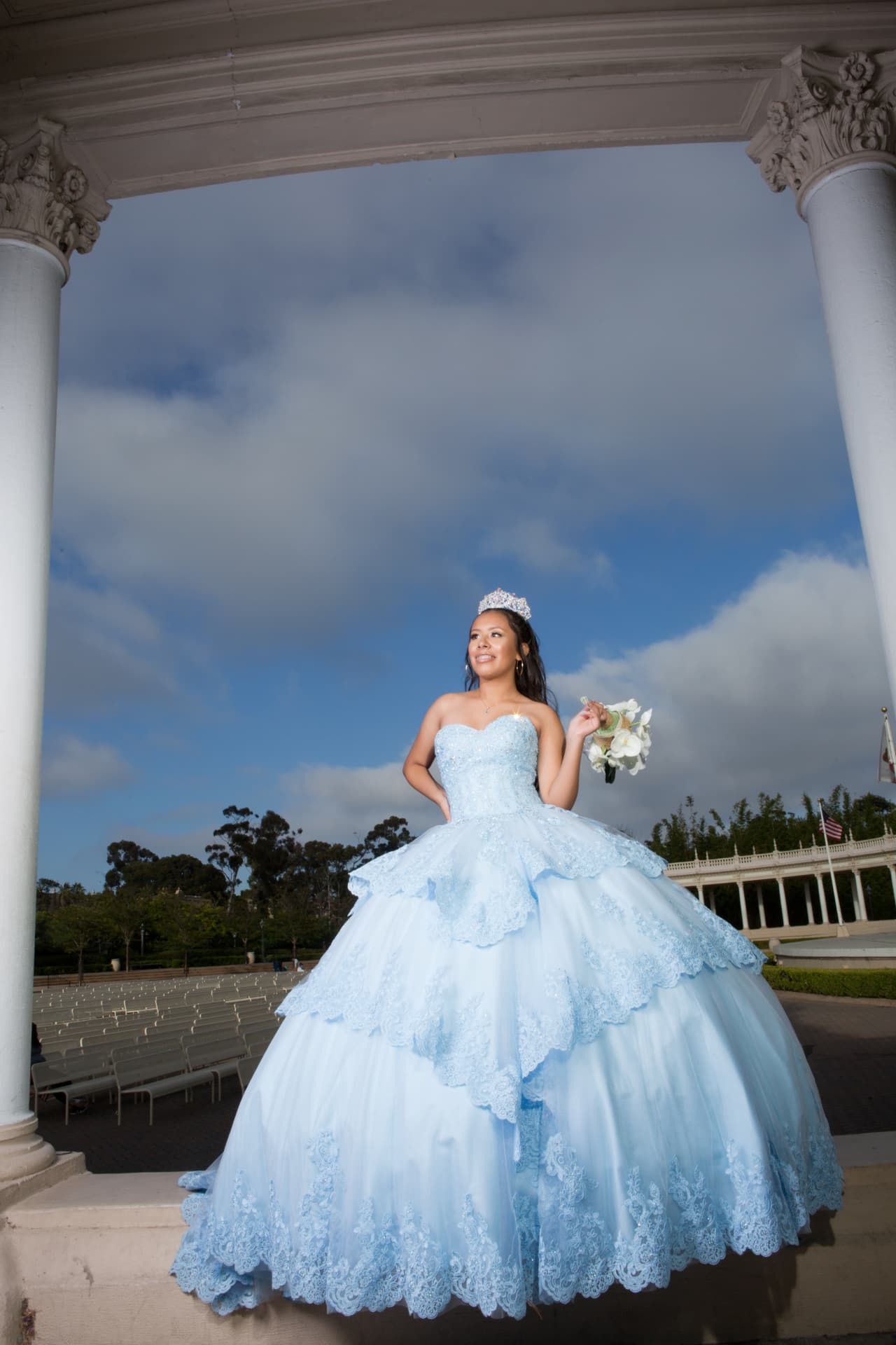 Quinceañera in a light blue lace gown and tiara holding a white bouquet beneath white columns