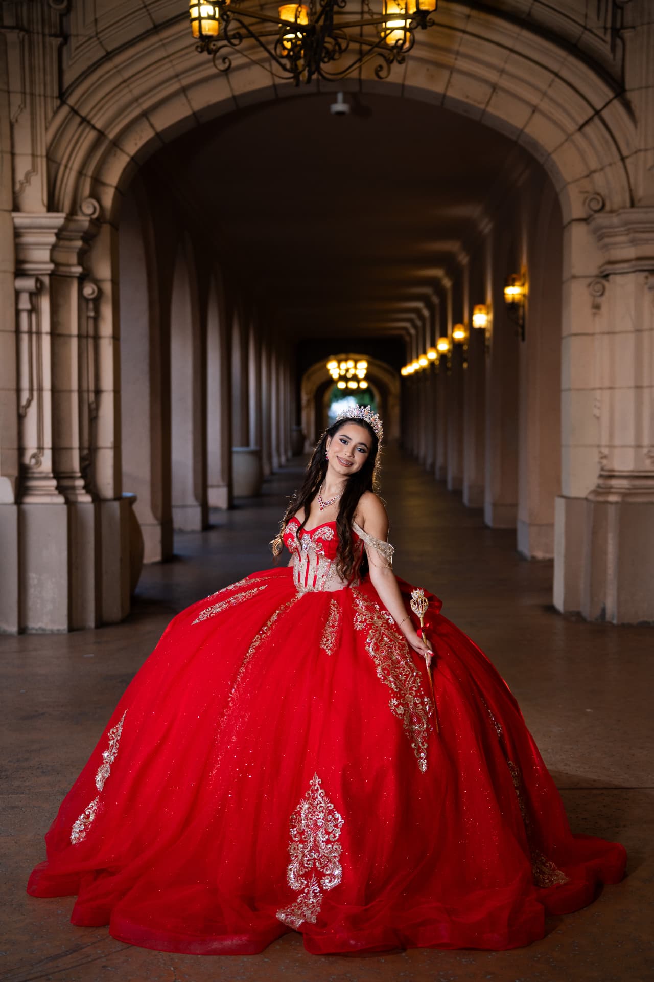 Quinceañera in a red gown with her golden scepter beneath the lanterns of a colonnade