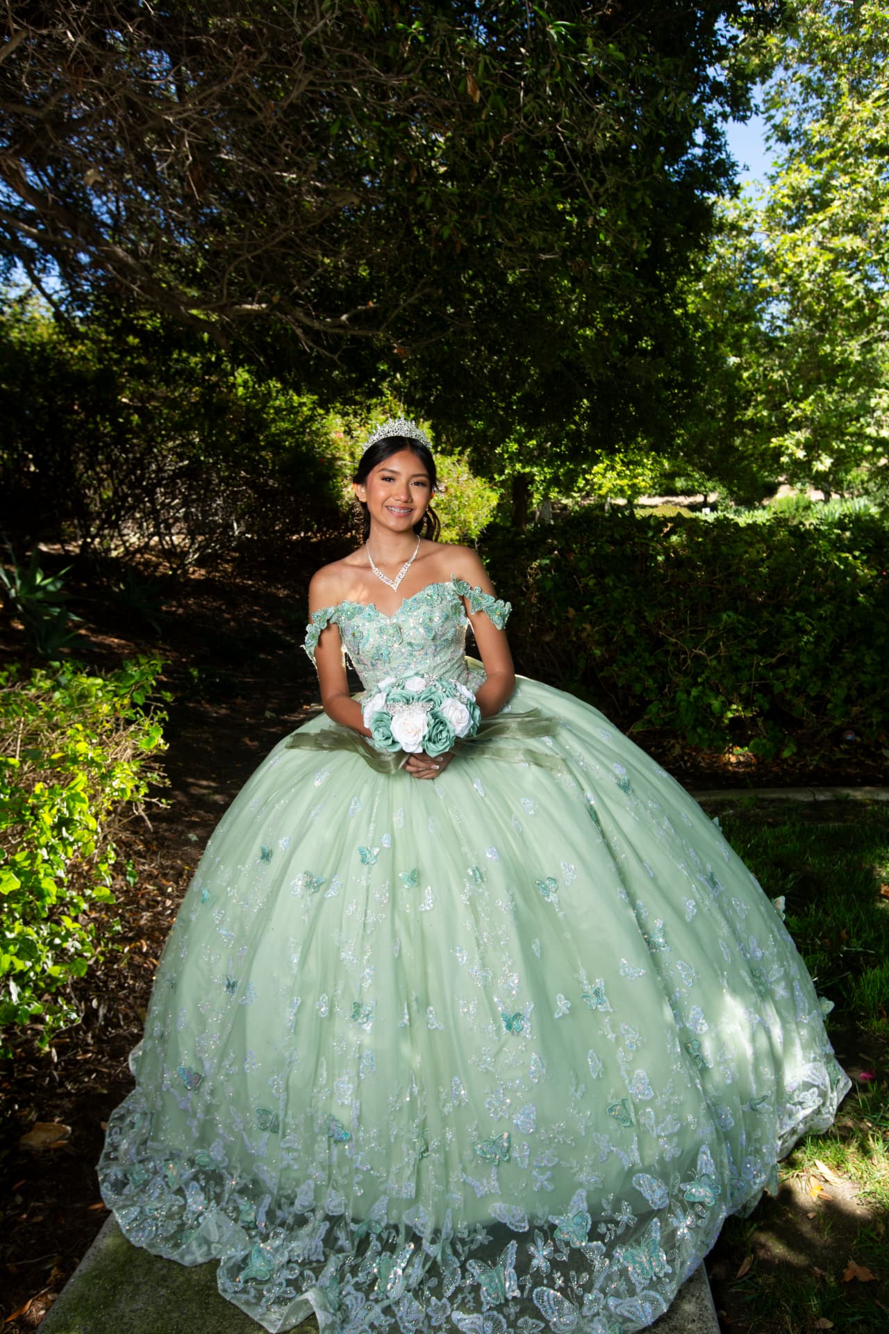 Quinceañera in a mint green butterfly gown holding her bouquet beneath the trees