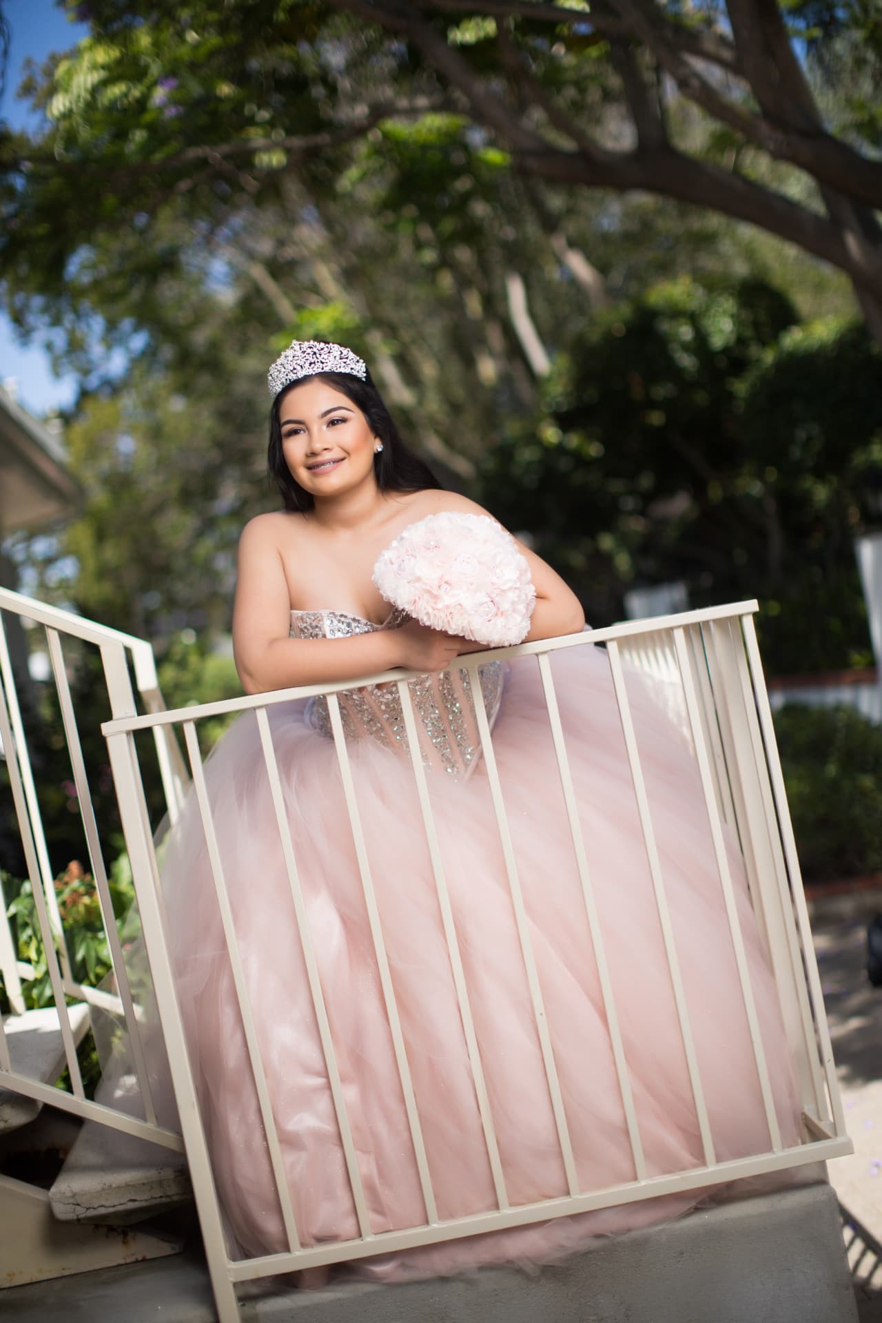 Quinceañera in a blush pink gown leaning on a white balcony railing with a pink bouquet