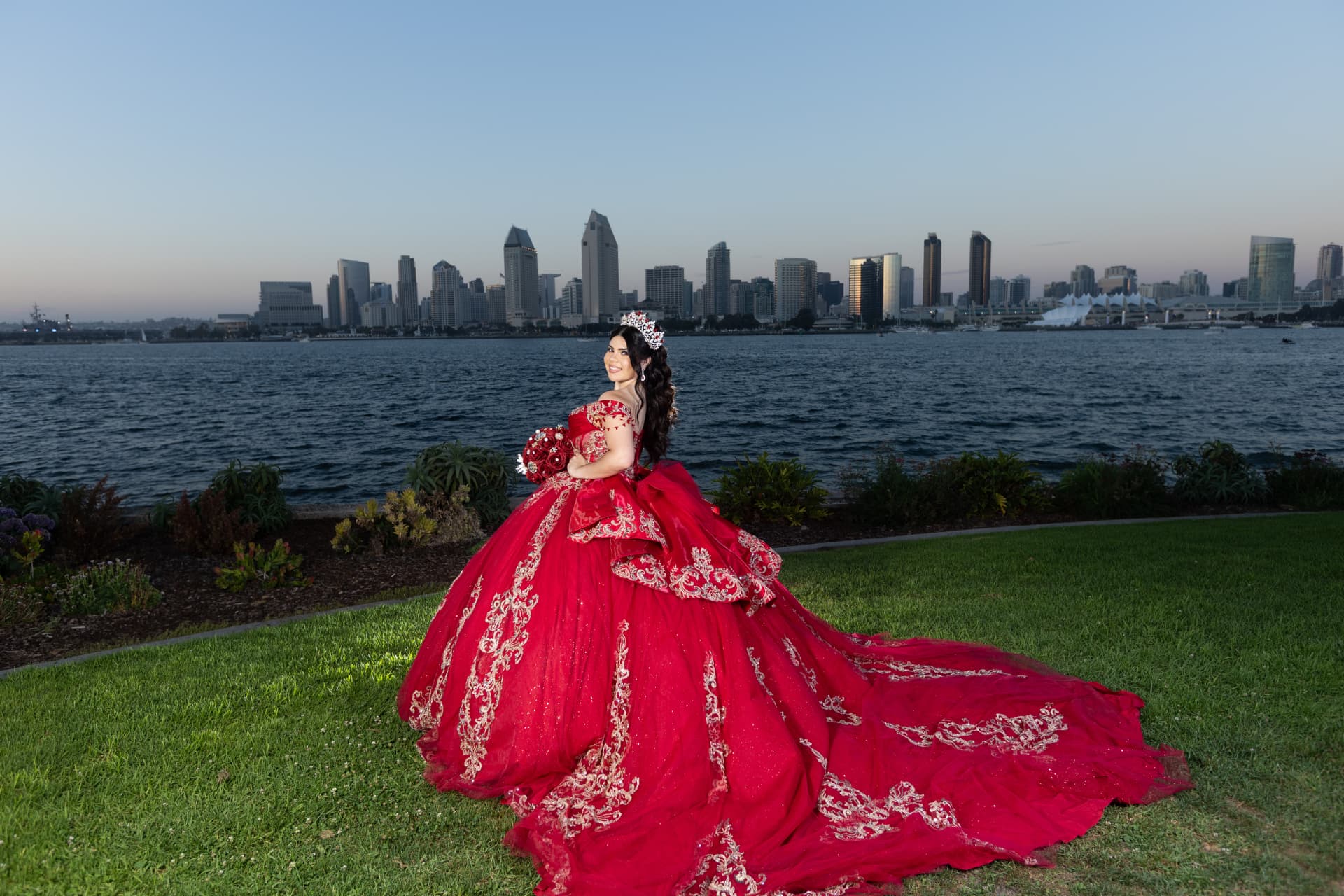 Quinceañera in a red gown with her train trailing across the grass before the waterfront skyline