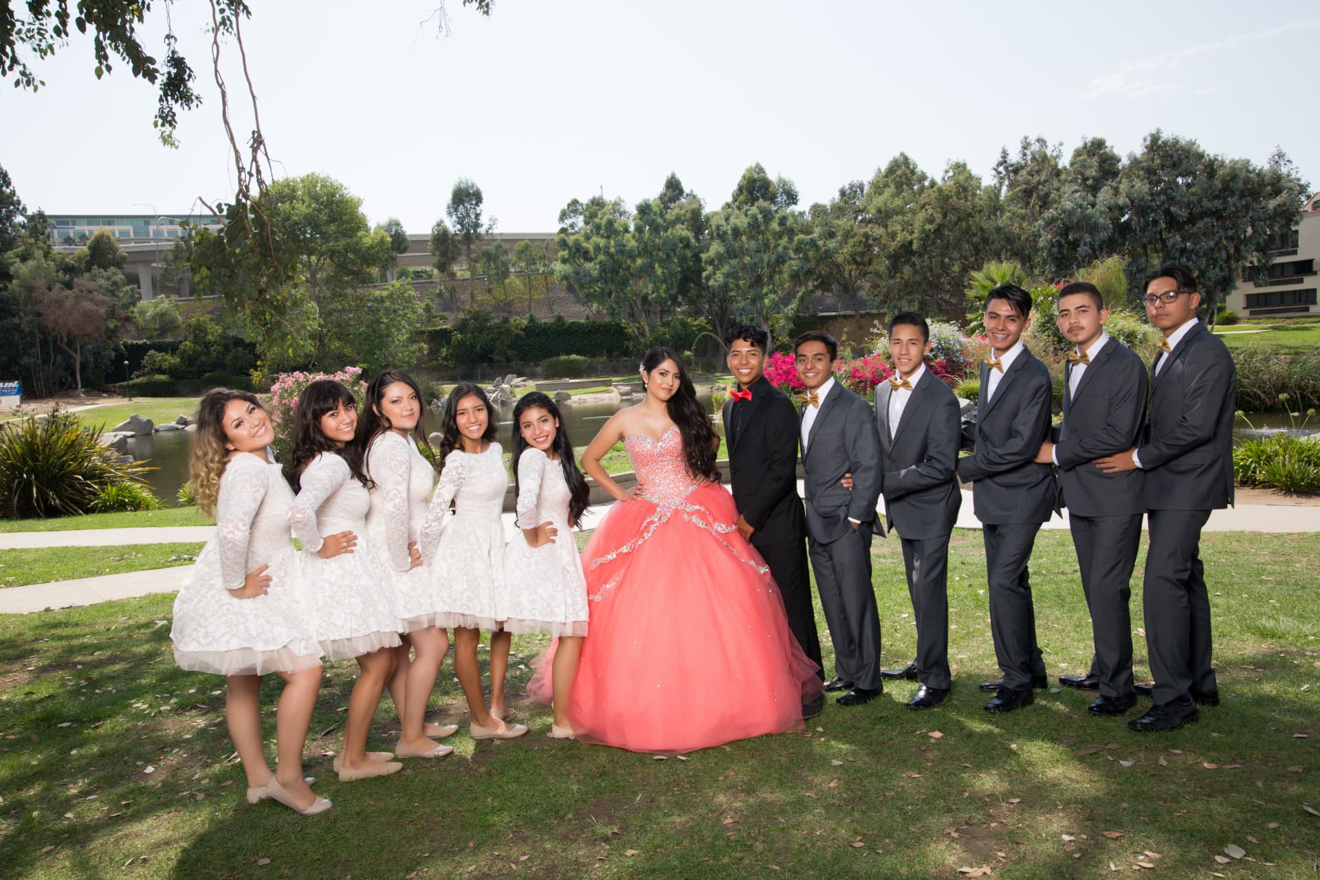 Quinceañera in a coral gown flanked by her damas in white lace dresses and chambelanes in gray suits in a park