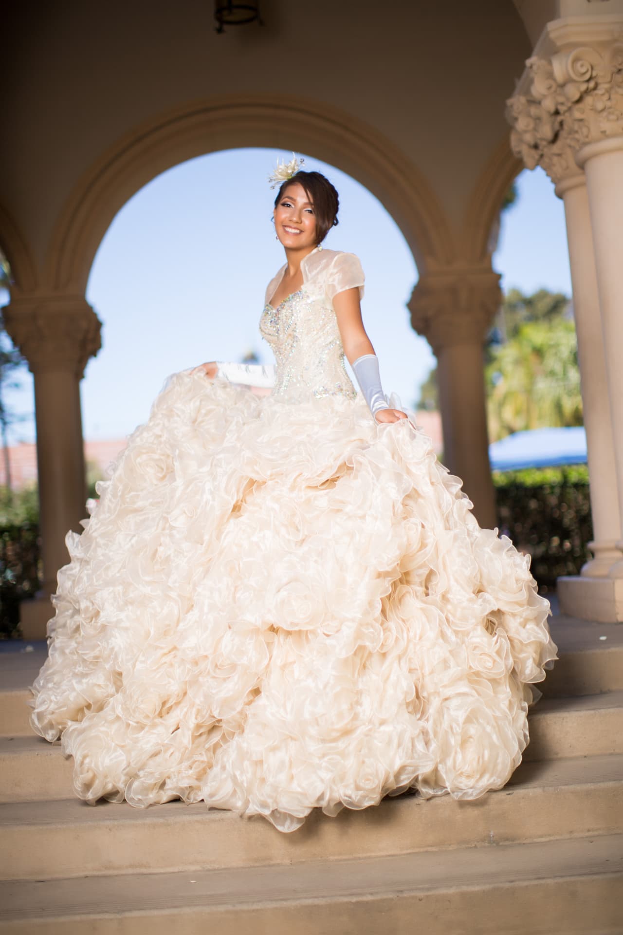 Quinceañera in an ivory rosette gown standing beneath arched colonnades