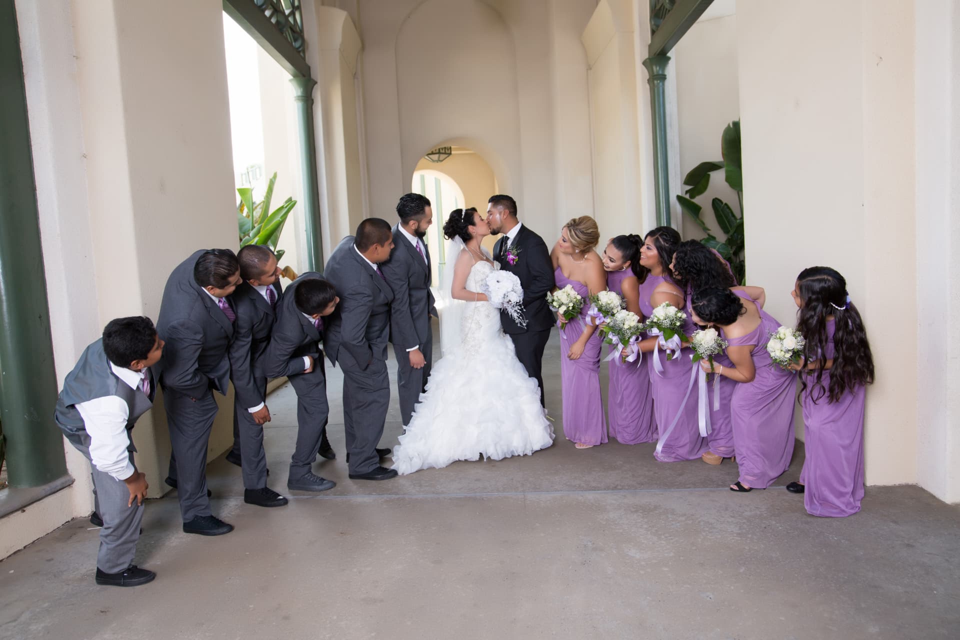 Wedding party leaning toward the newlyweds as the bride and groom kiss beneath an archway