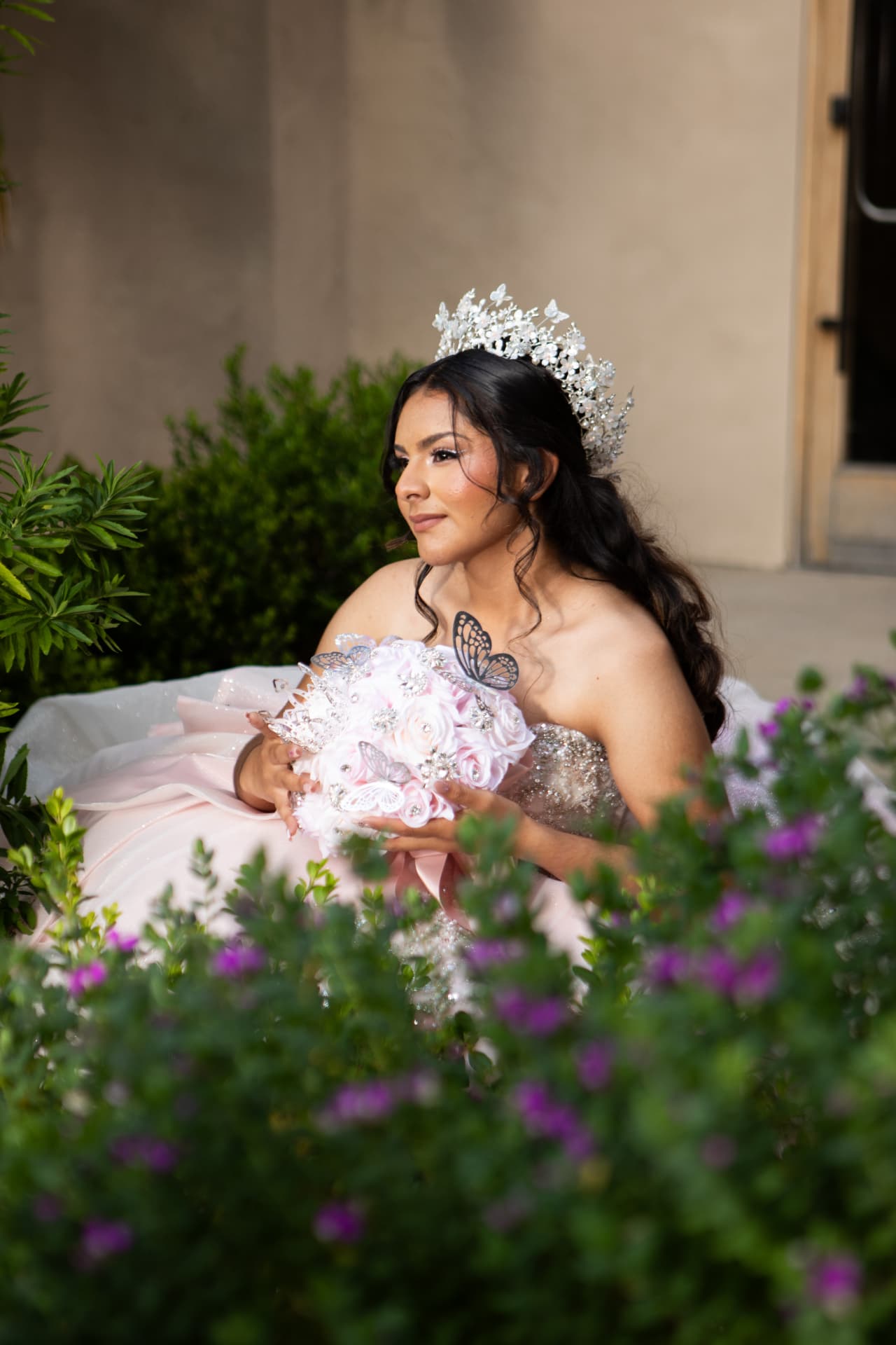 Quinceañera in a pink gown with a butterfly bouquet seated among flowering bushes