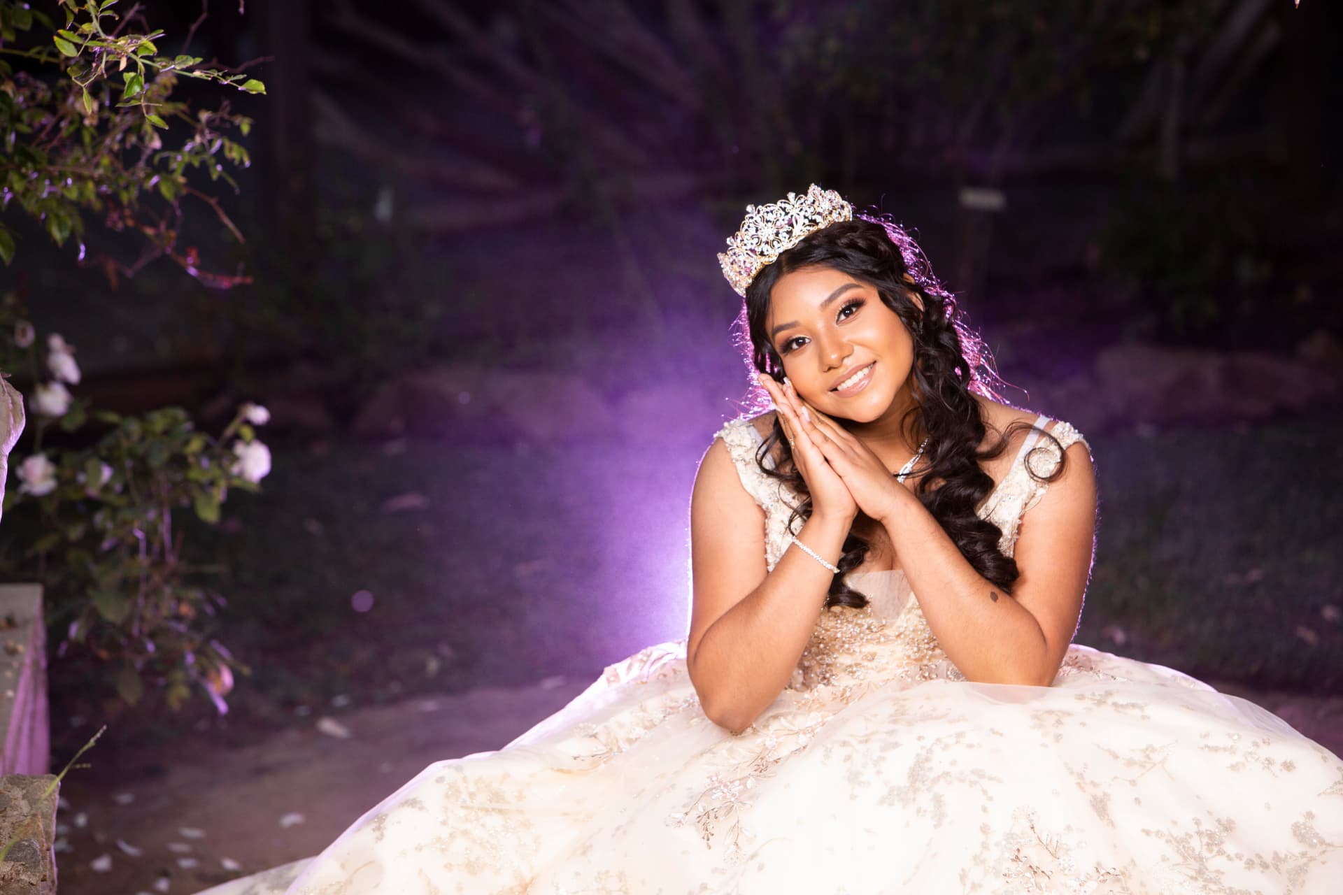 Quinceañera in a cream and gold gown with her hands together beneath a glowing crown and purple light