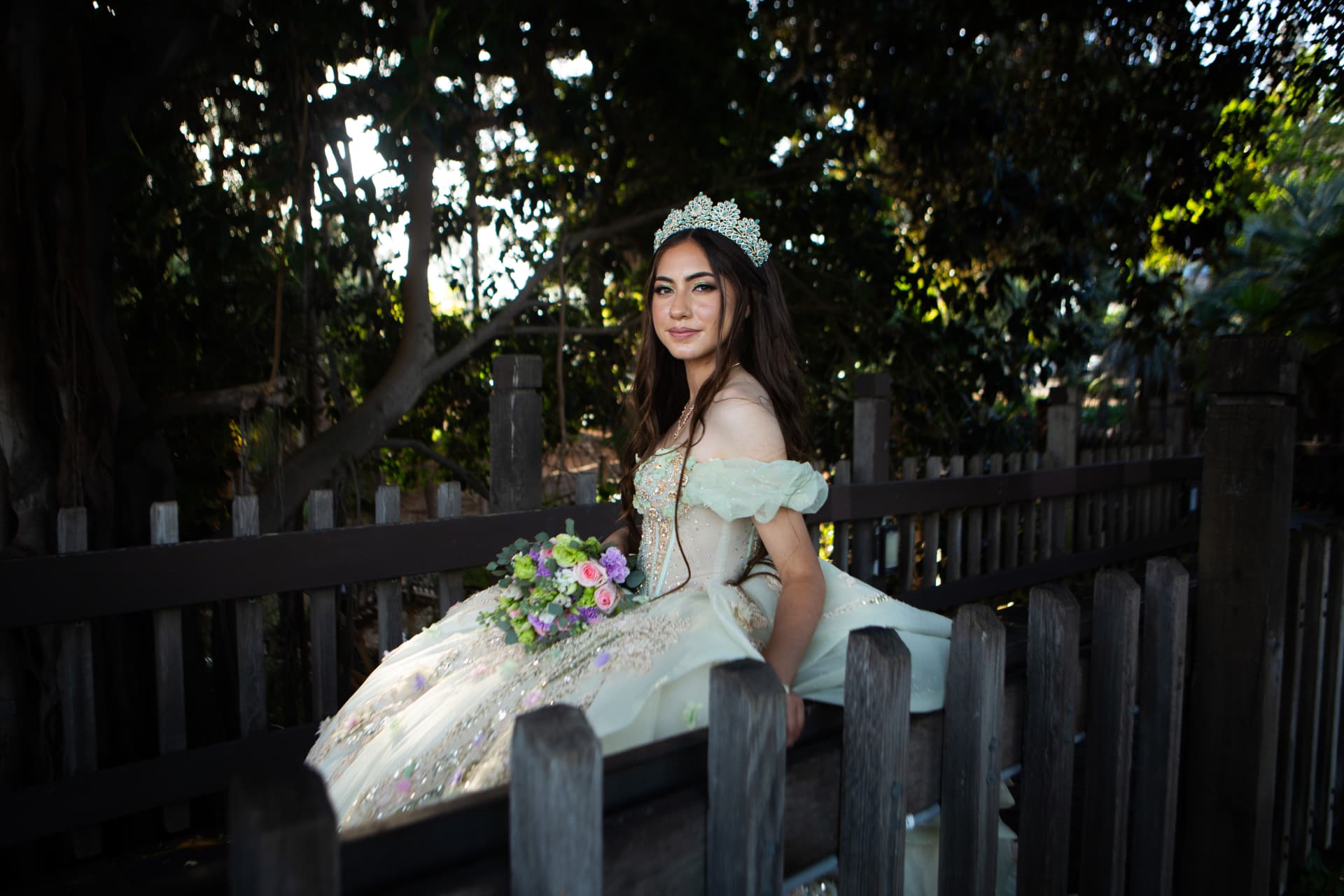 Quinceañera in a pastel gown seated on a wooden bridge holding her bouquet