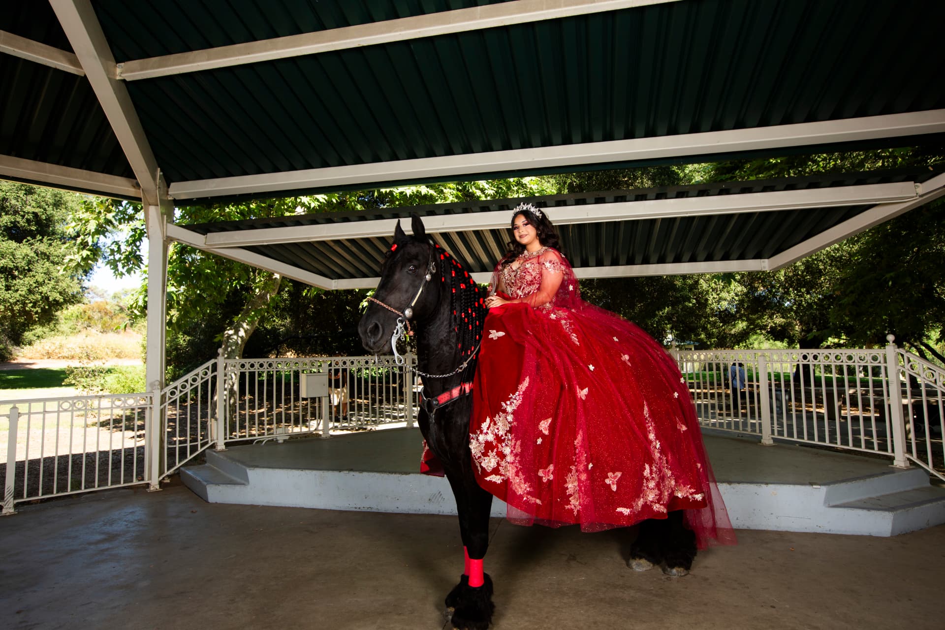 Quinceañera in a red gown seated on a black horse beneath a park pavilion