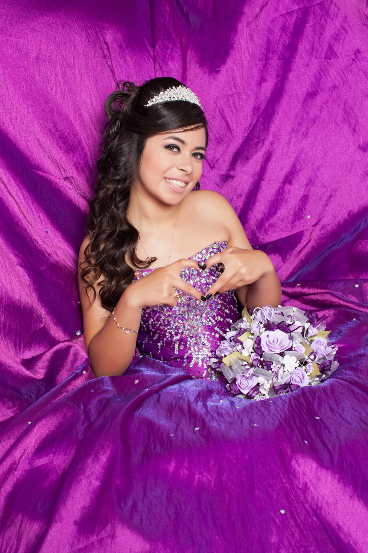 Quinceañera in a purple sequined gown making a heart with her hands on a purple backdrop