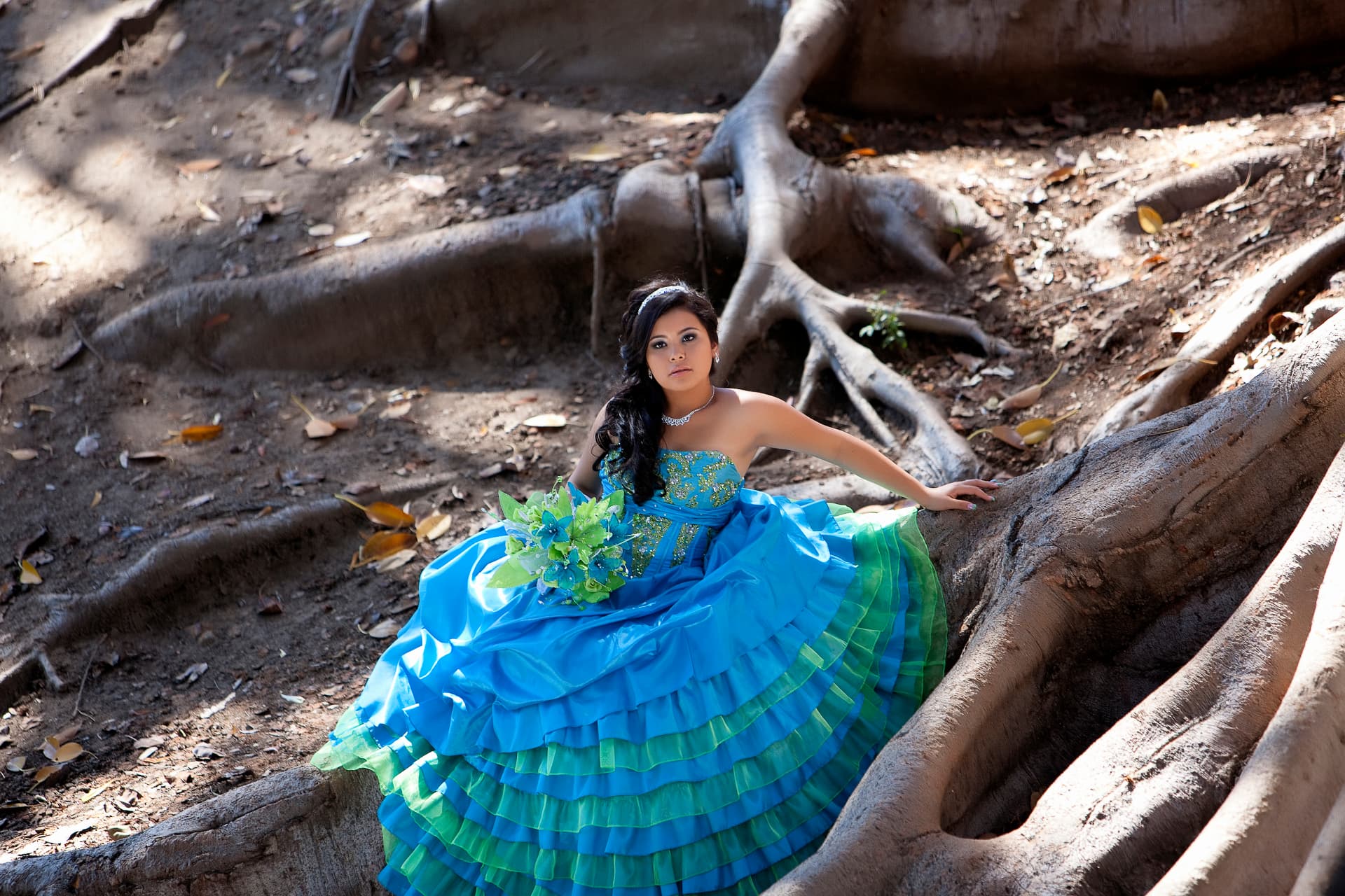 Quinceañera in a blue and green ruffled gown posed among sprawling tree roots