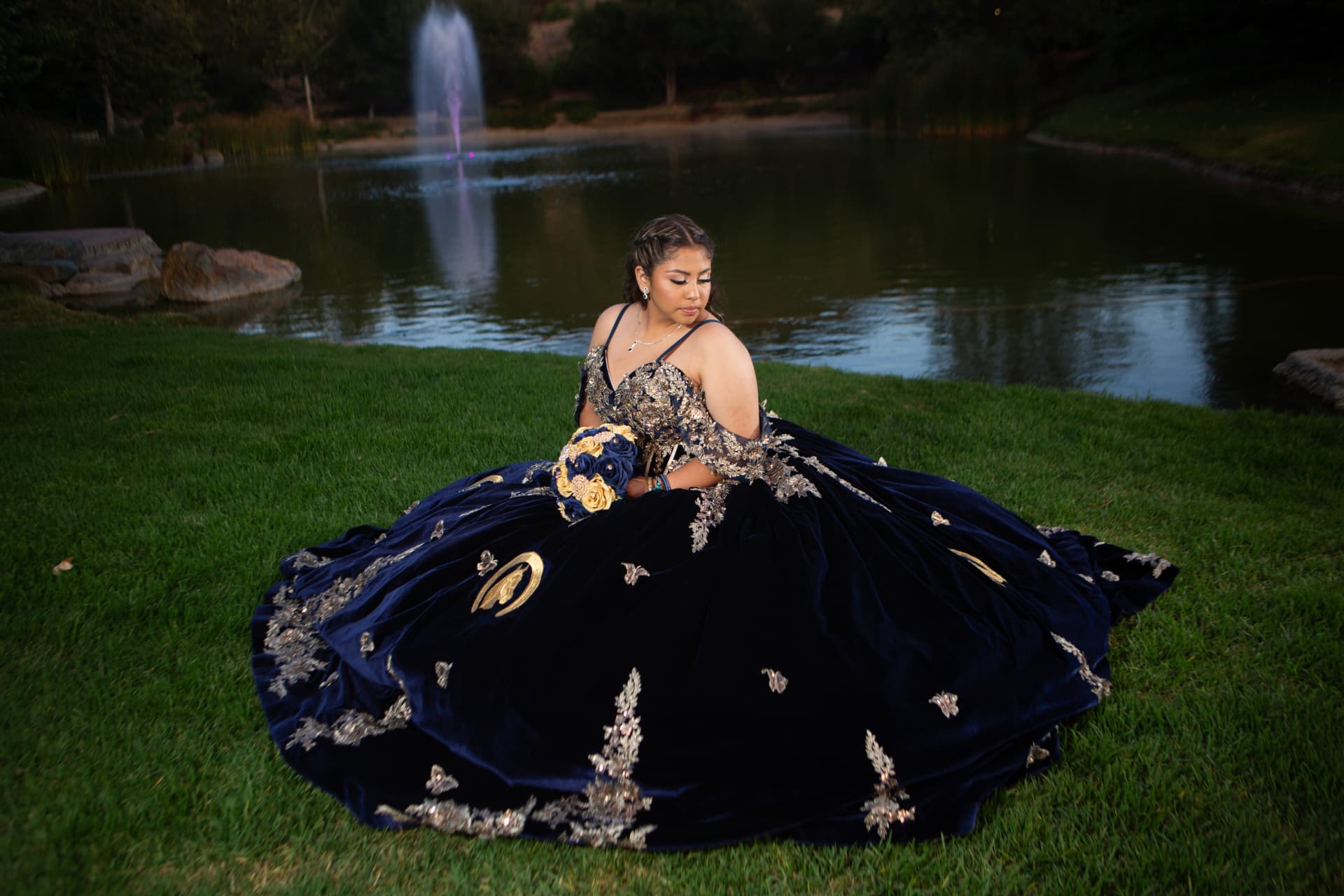 Quinceañera in a navy and gold gown seated on the grass beside a lit fountain at dusk