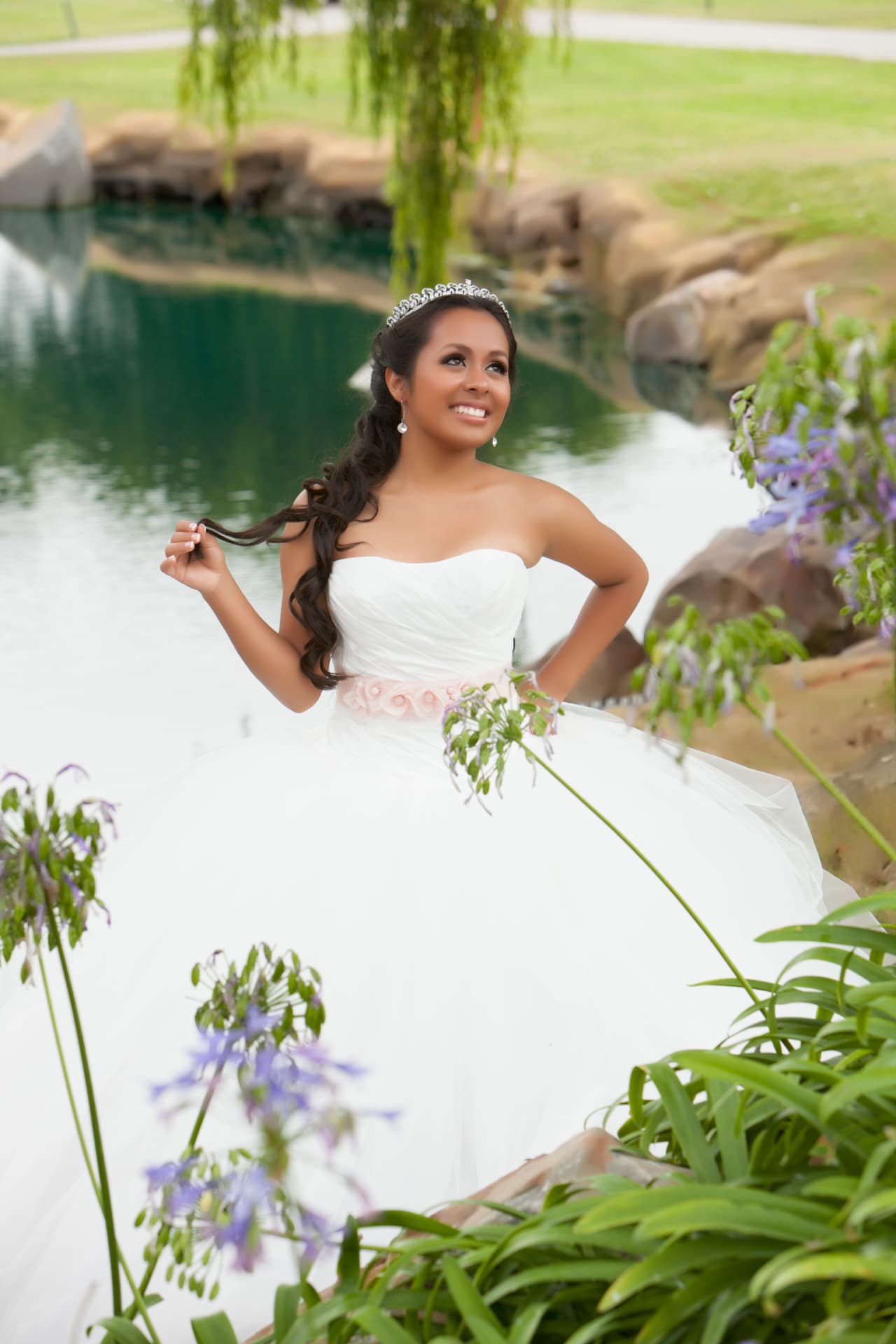 Smiling quinceañera in a white gown with a pink sash beside a pond framed by purple flowers