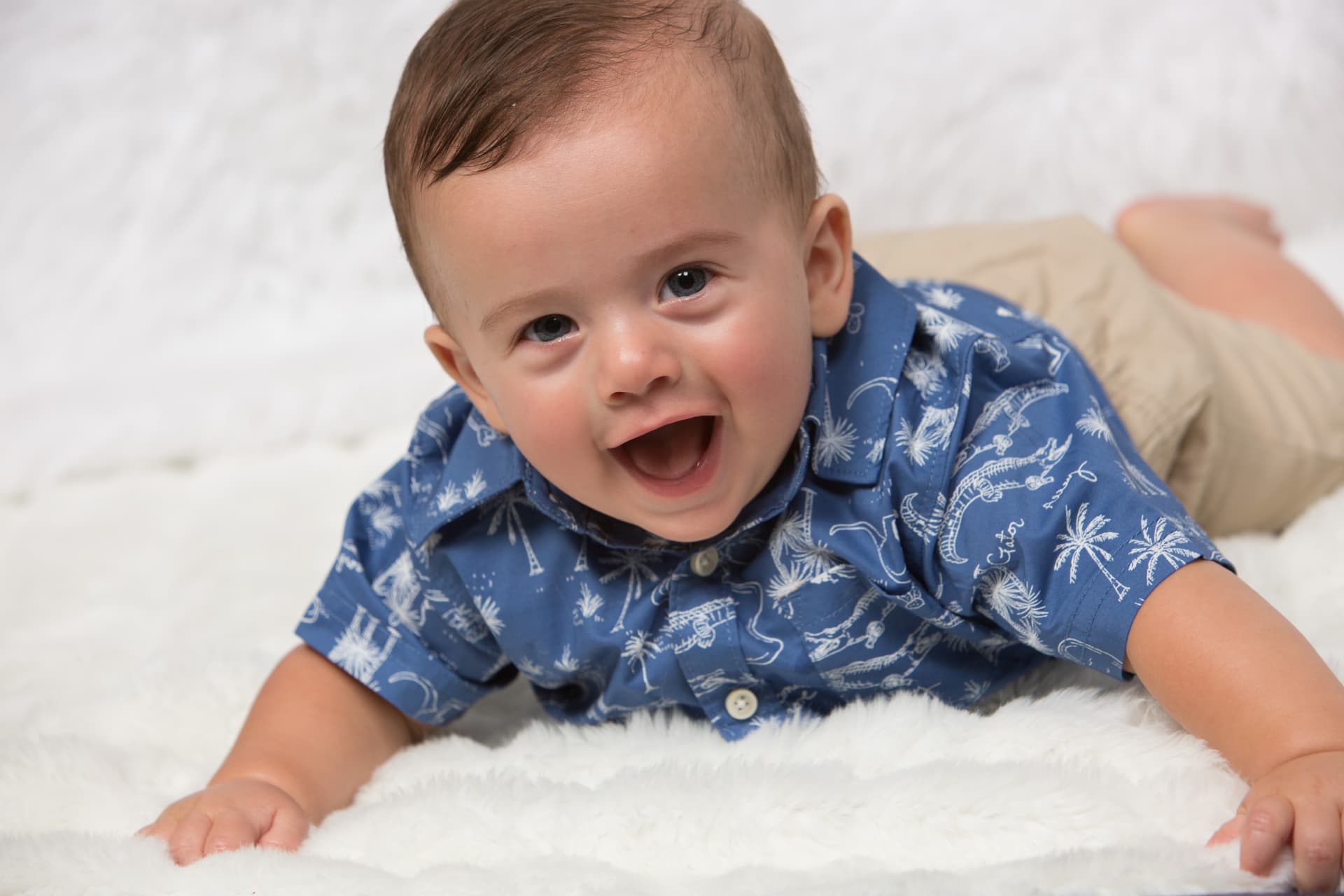 Laughing baby boy in a blue patterned shirt on a soft white blanket