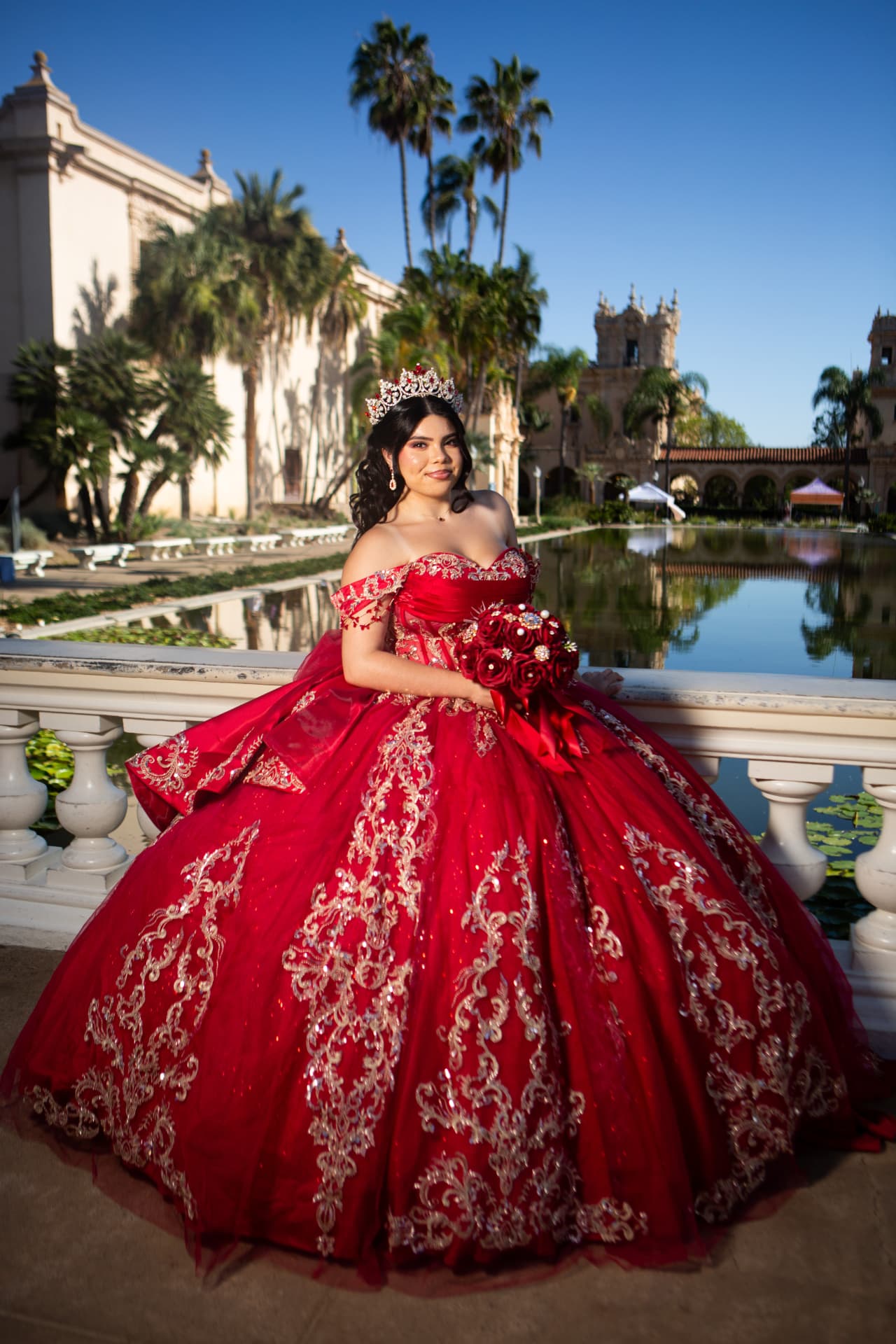 Quinceañera in a red gown by a reflecting pond with palms and the Balboa Park tower behind her