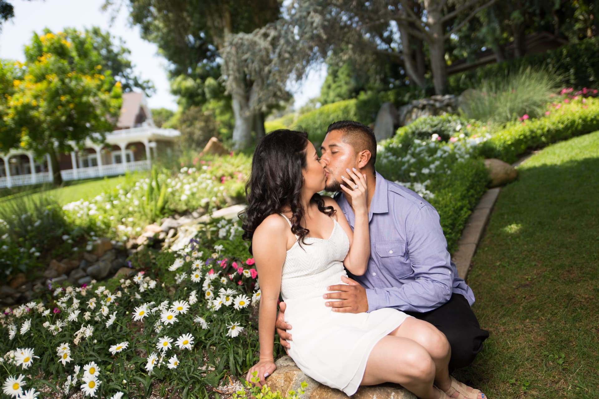Engaged couple kissing beside white flowers in a sunlit garden