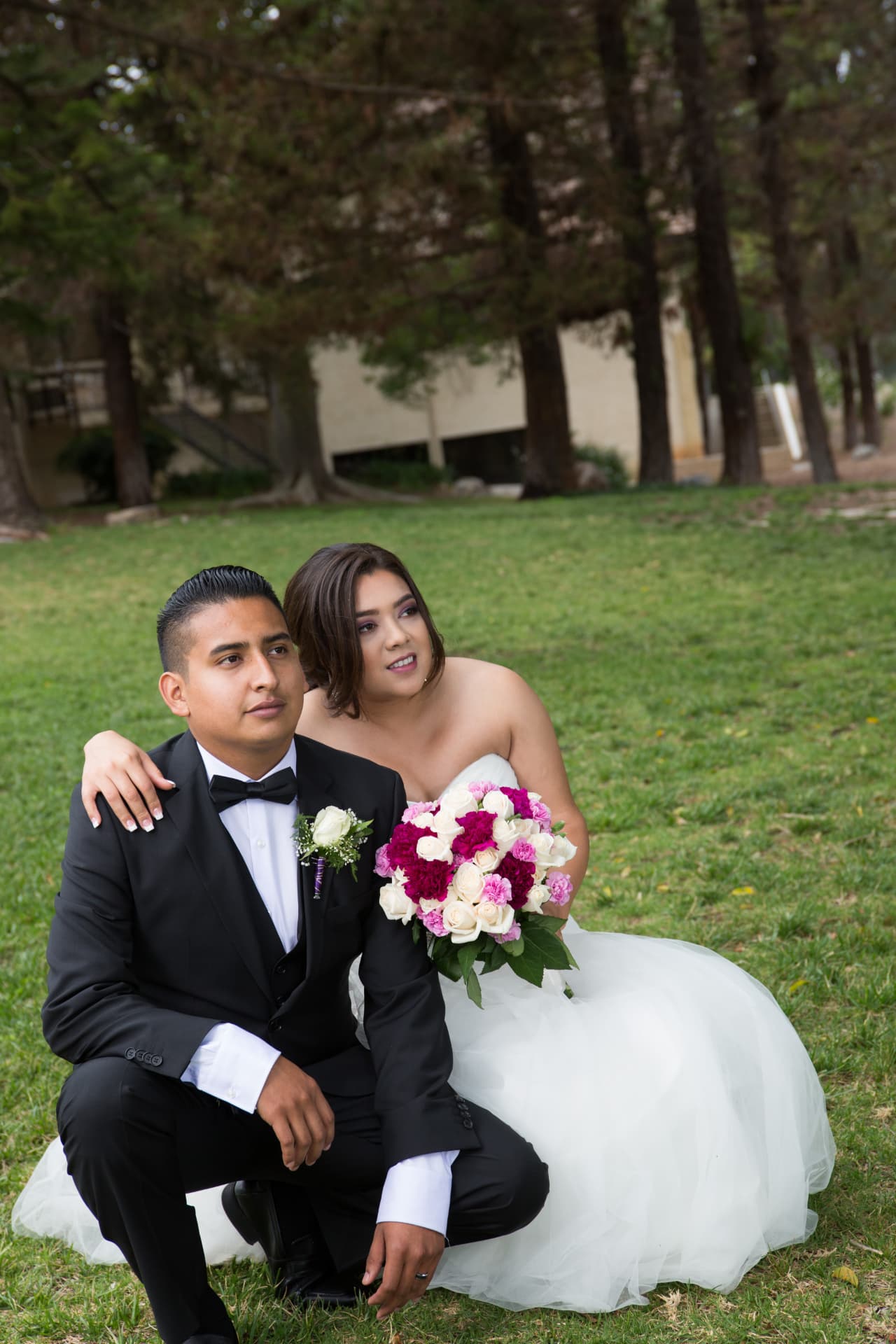 Bride with a pink and white bouquet embracing her seated groom on a green lawn