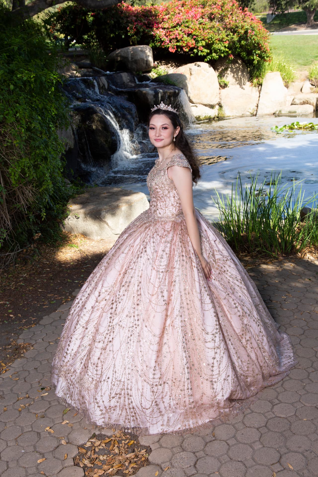 Quinceañera in a rose gold embroidered gown beside a pond with a waterfall and greenery