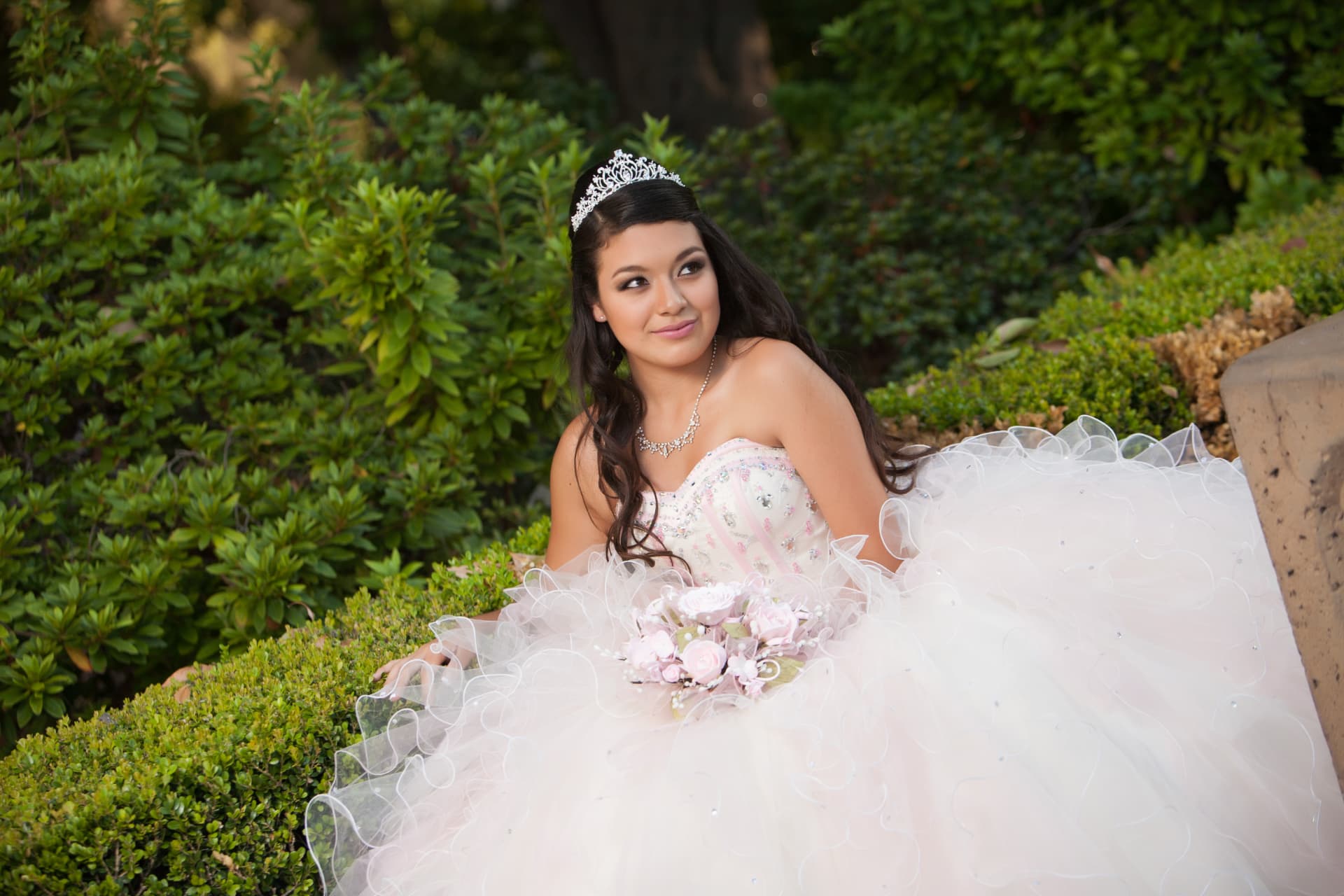 Quinceañera in a blush gown with a pink bouquet and tiara seated among green hedges