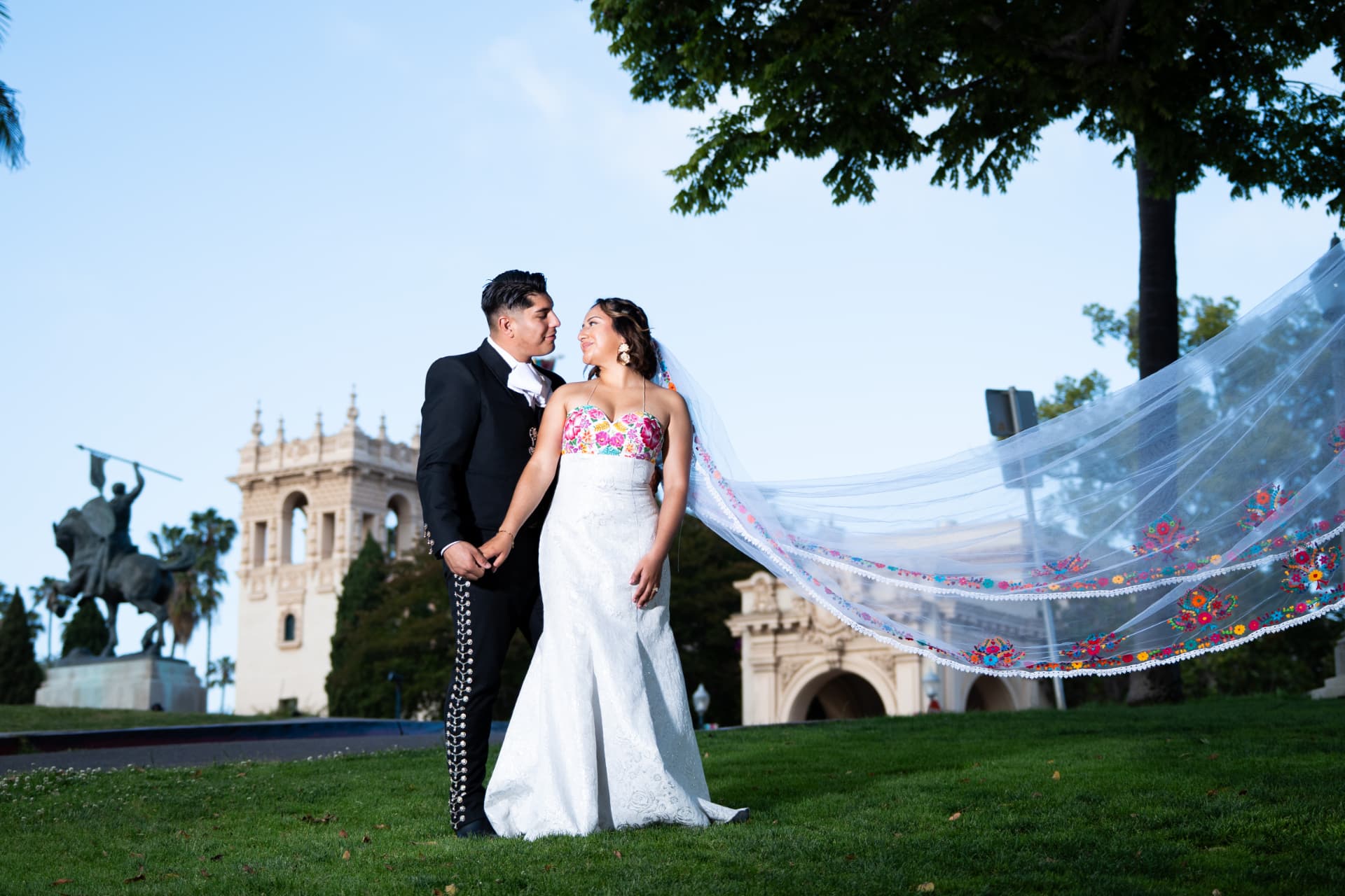 Bride and groom standing before historic architecture with her embroidered veil flowing behind them