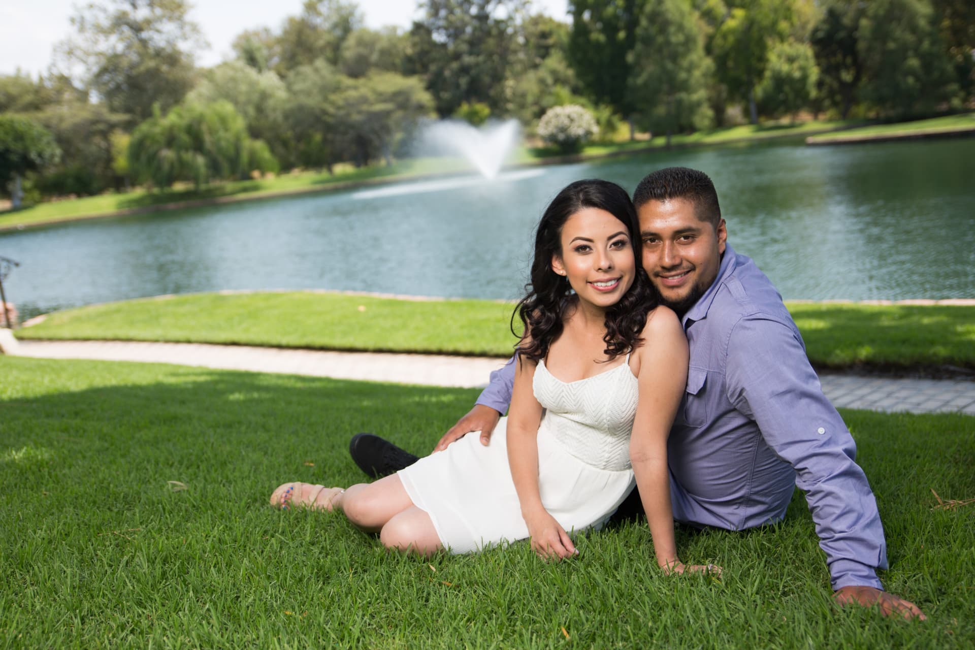 Engaged couple seated together on a lakeside lawn with a fountain behind them