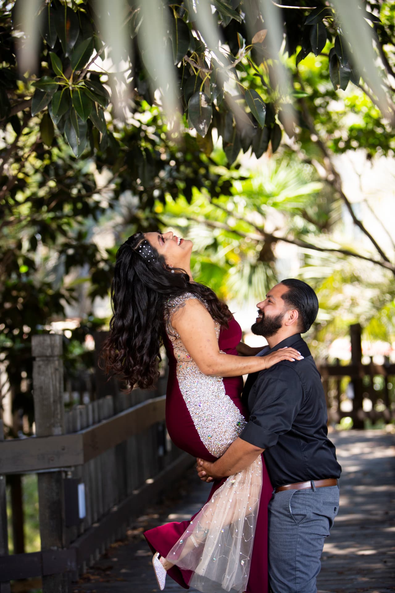 Engaged couple laughing during a playful lift beneath garden trees