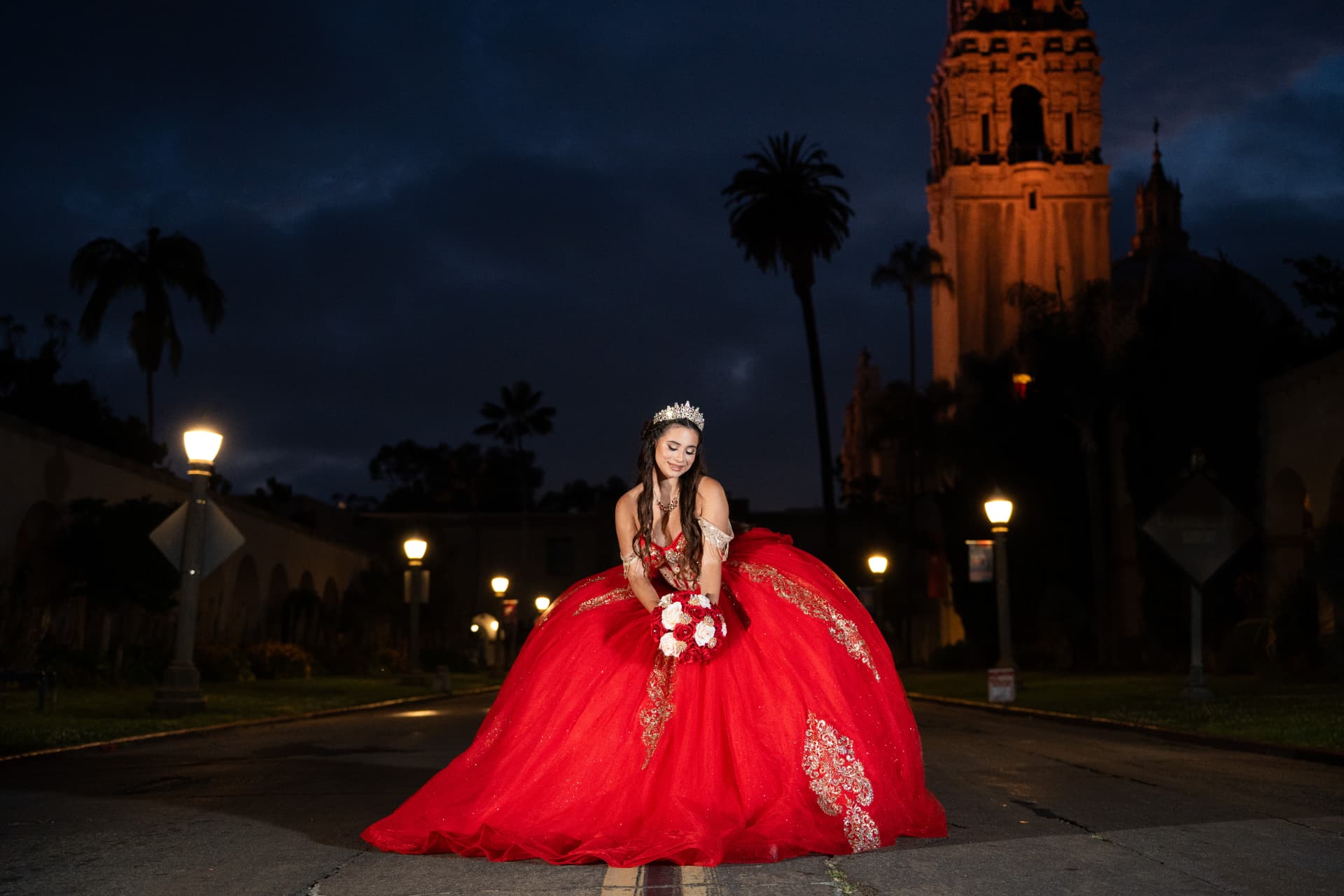 Quinceañera in a red gown holding her bouquet before the illuminated Balboa Park tower at night