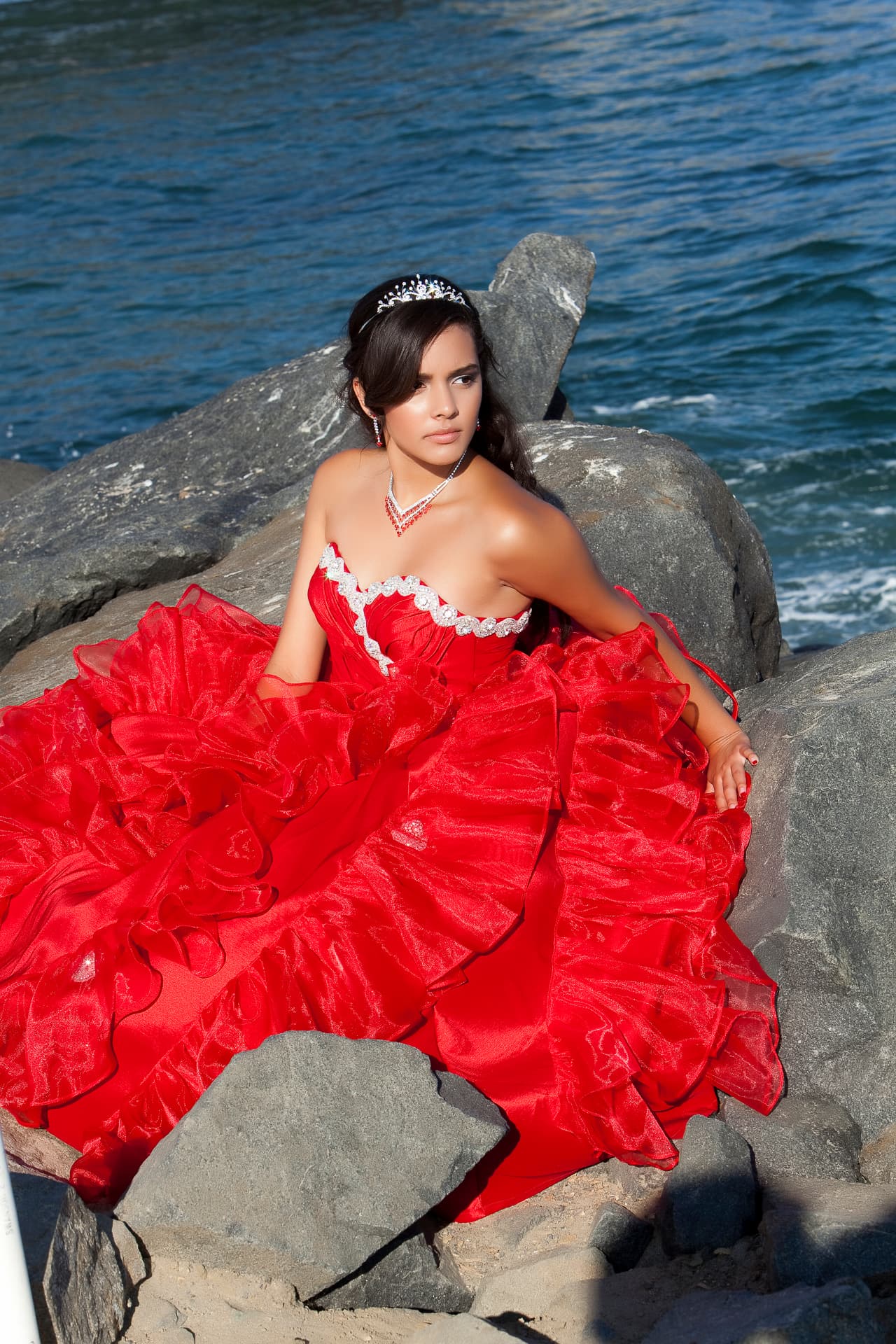 Quinceañera in a red ruffled gown with a tiara seated among dark breakwater rocks by the ocean