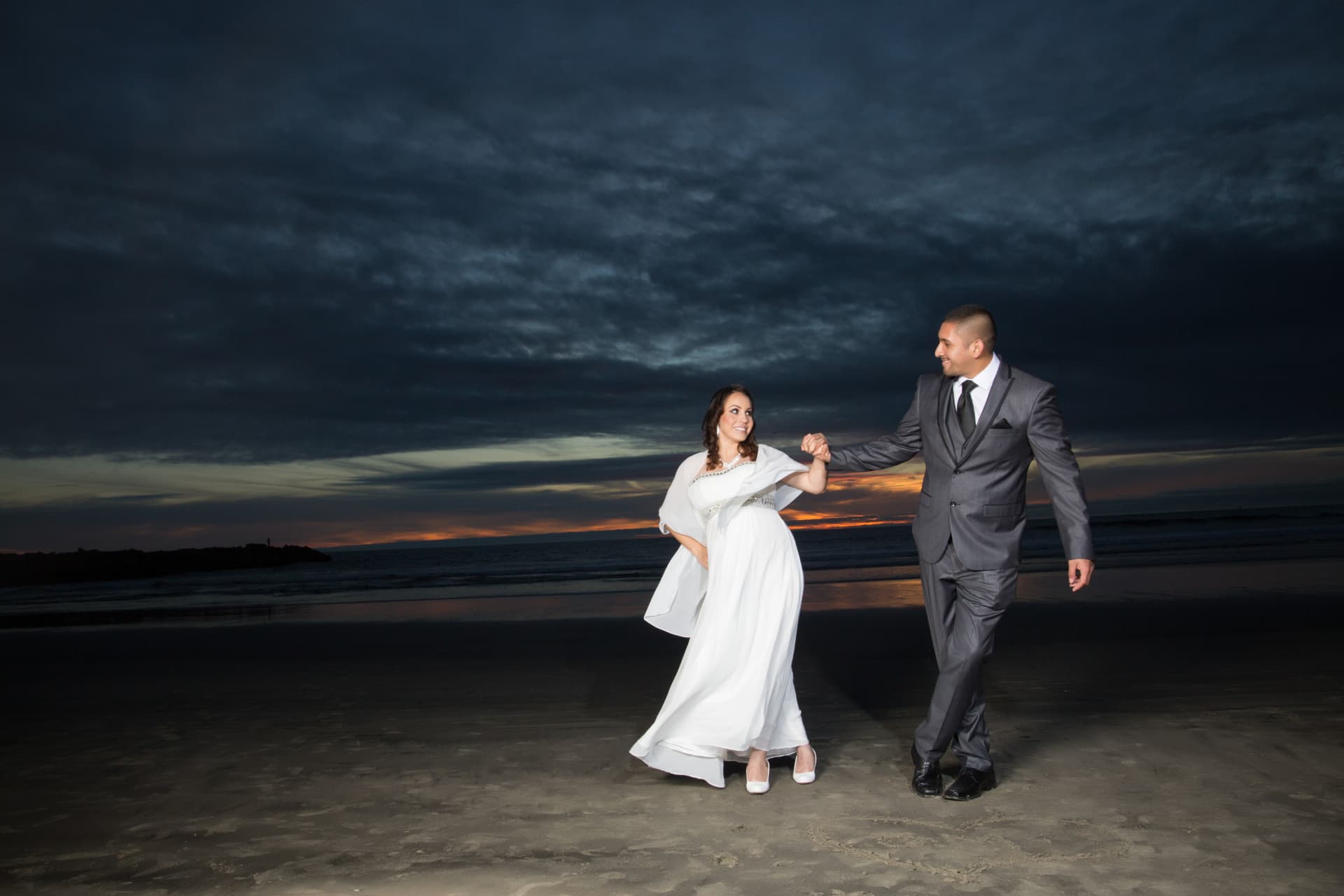 Newlywed couple walking hand in hand on the beach beneath a dramatic sunset sky