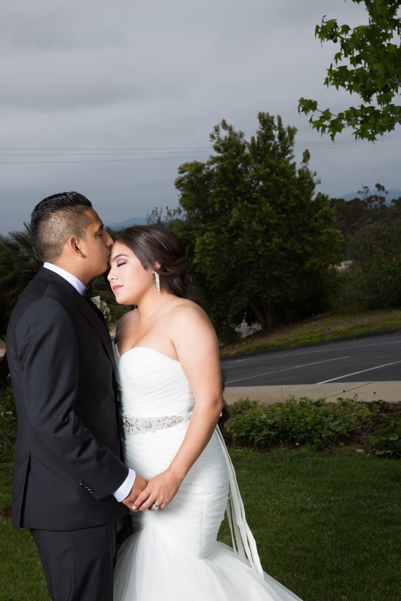 Groom kissing his bride’s forehead as they hold hands outdoors
