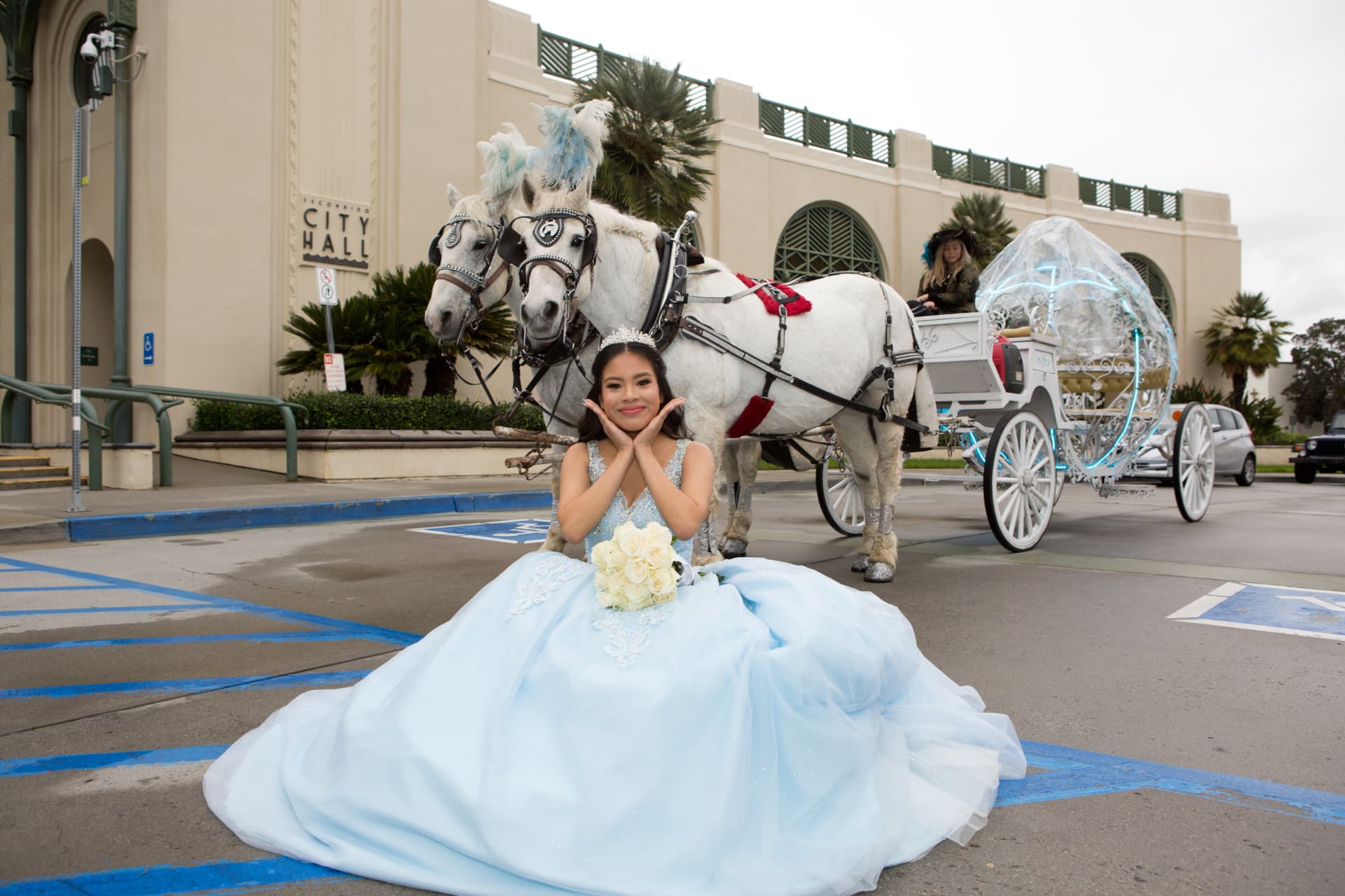 Quinceañera in a light blue gown kneeling before white horses and a glass carriage at city hall