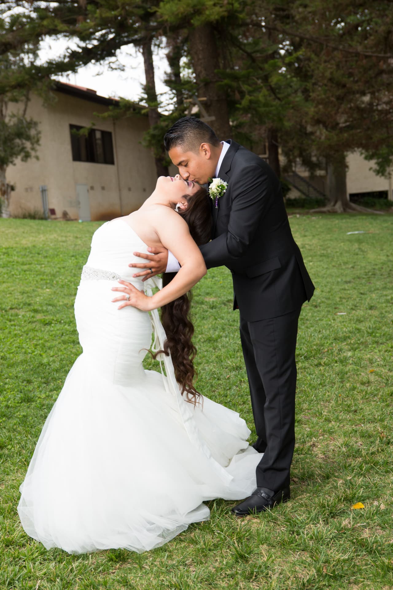Groom dipping and kissing his bride on a green lawn beneath tall trees