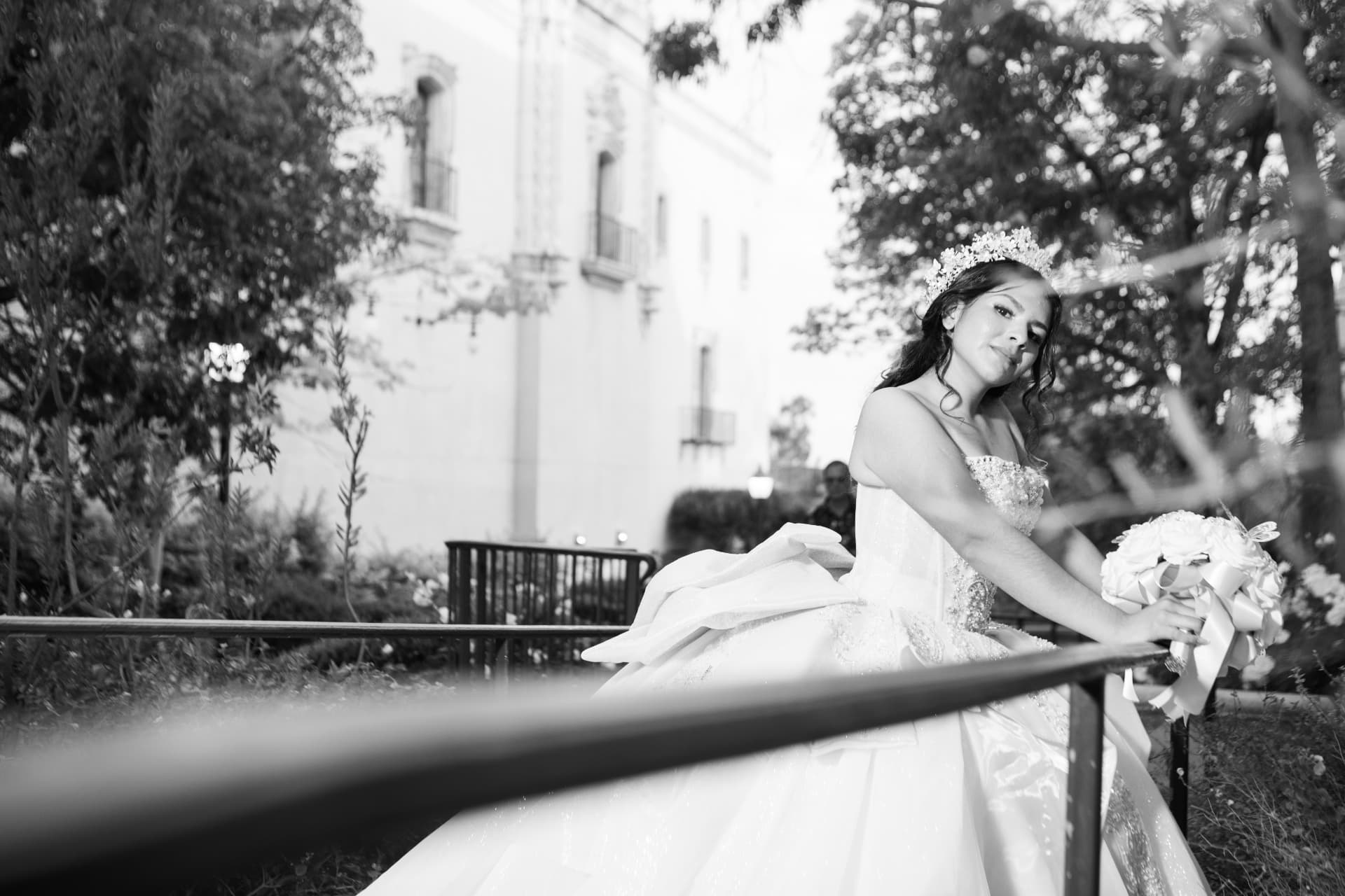 Black and white photo of a quinceañera in a white gown leaning on a garden railing with her bouquet
