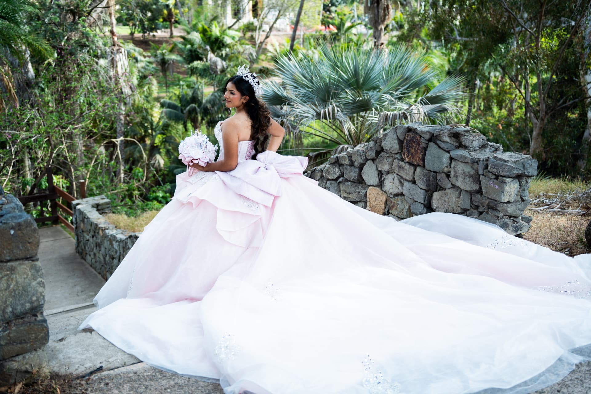 Quinceañera in a pink gown with a large bow looking back as her long train spreads down a stone garden path