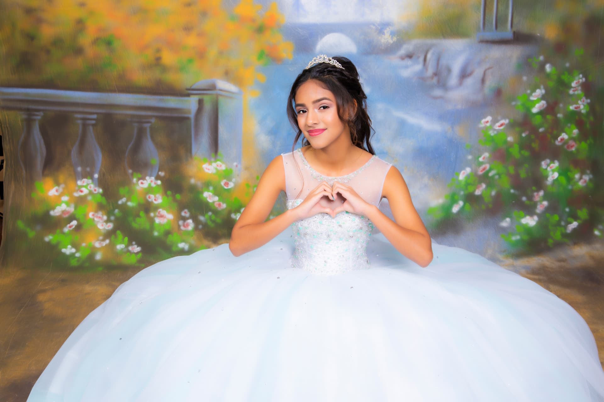 Quinceañera in a white gown making a heart with her hands before a painted lakeside backdrop