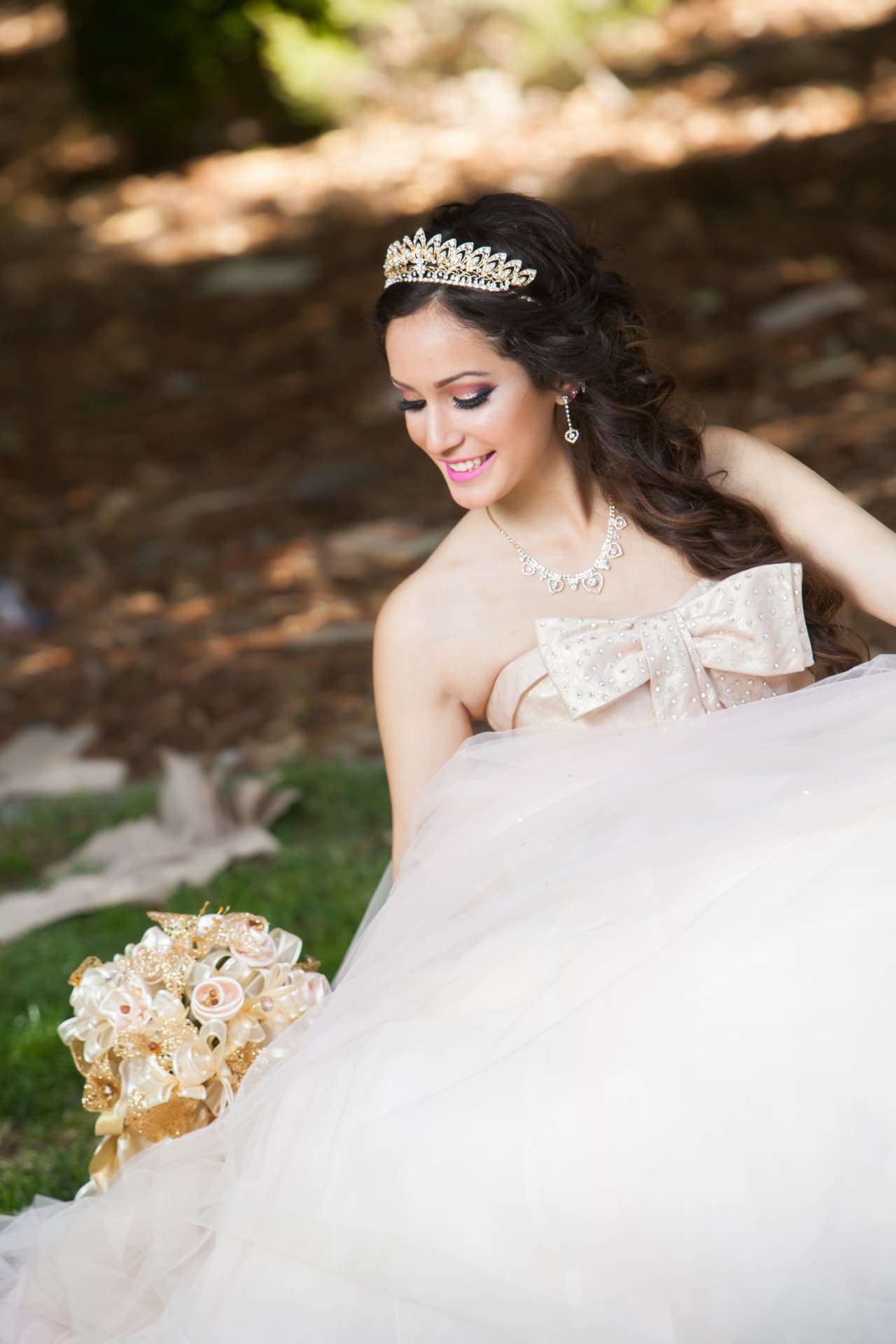 Quinceañera in a champagne gown with a jeweled bow and gold tiara smiling in a sunlit garden