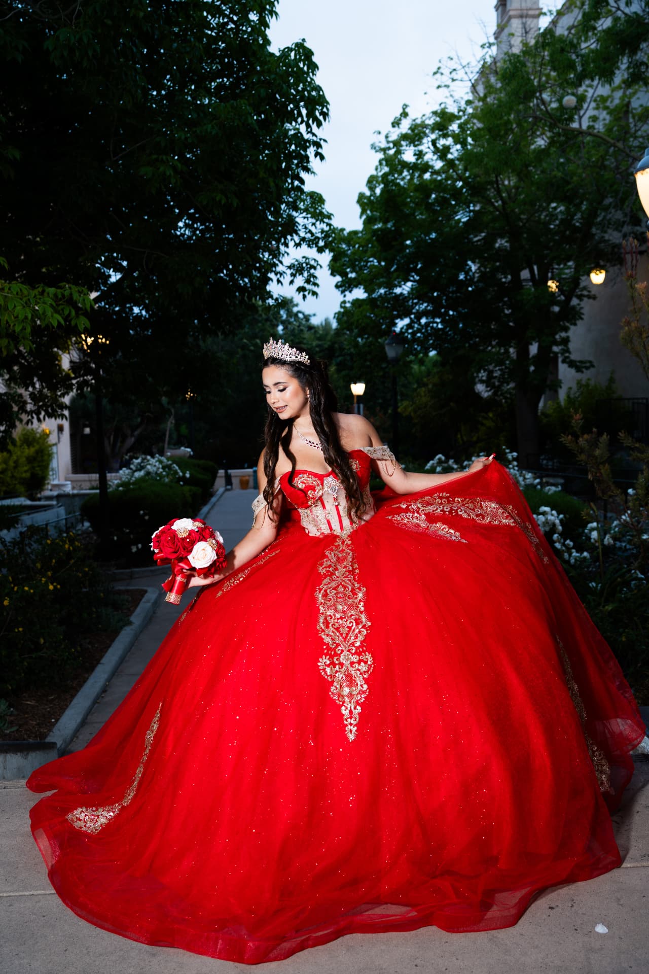 Quinceañera in a red gown spreading her skirt as she holds her bouquet in a garden at dusk