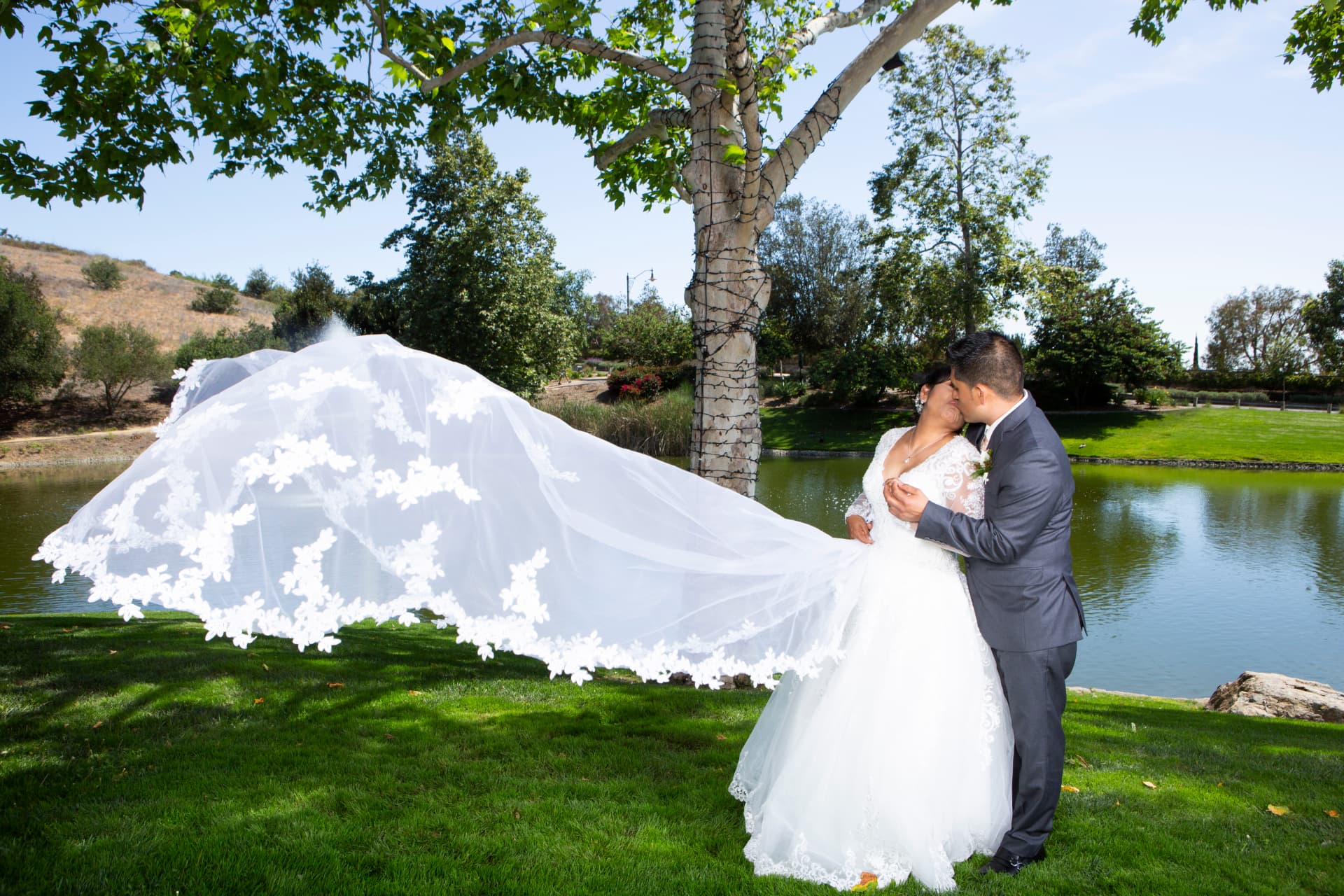 Bride and groom kissing beside a lake as her lace veil sweeps across the lawn