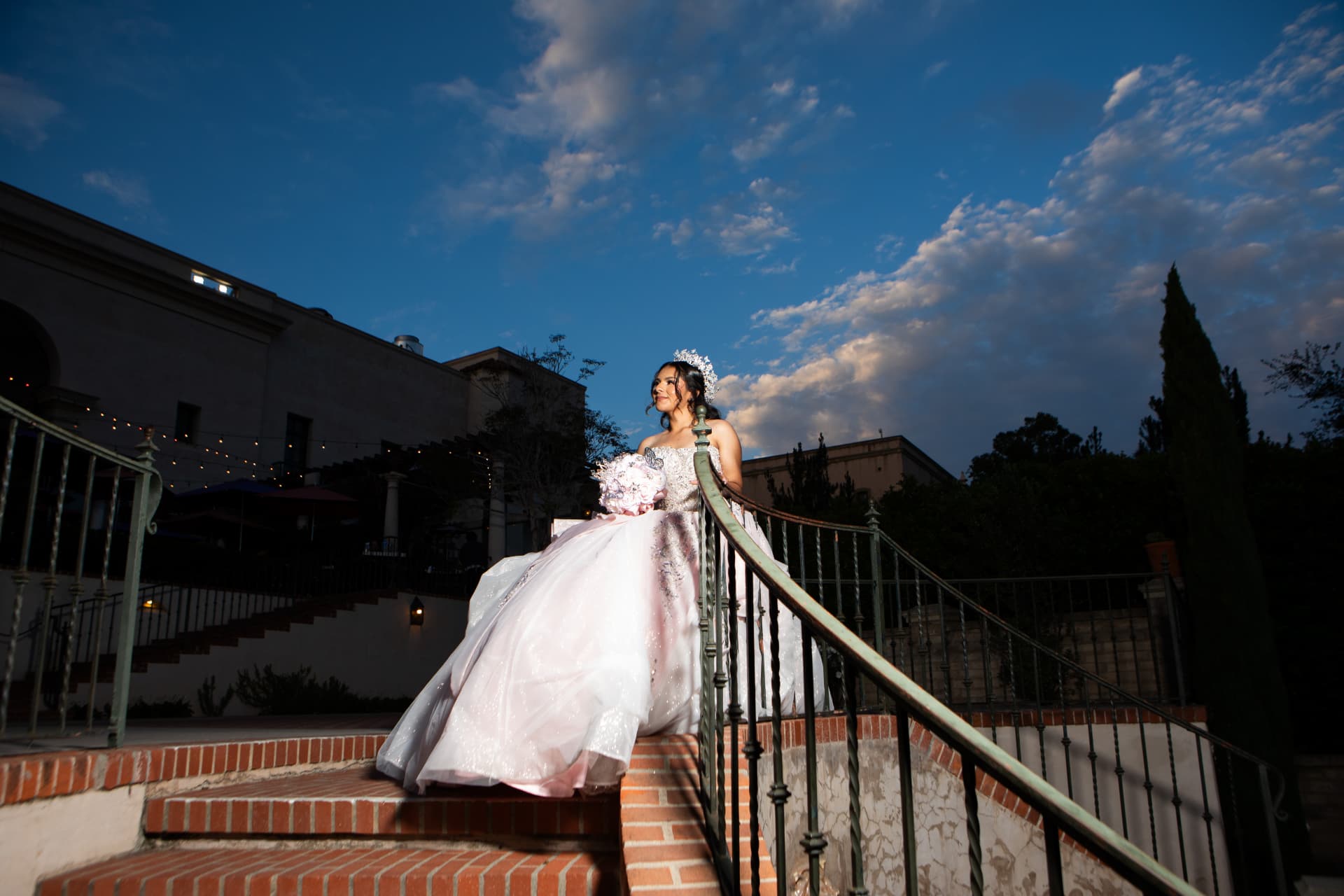 Quinceañera in a pink gown on brick steps beneath string lights and a blue dusk sky