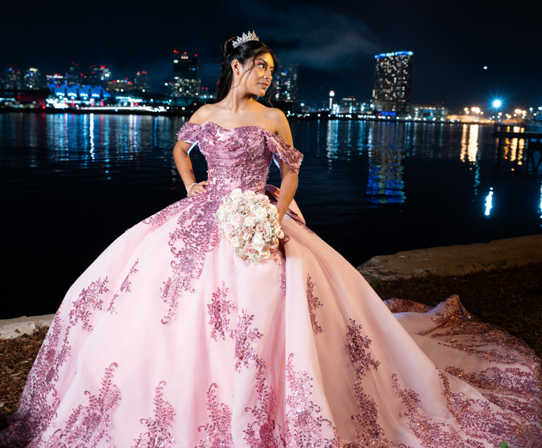 Quinceañera in a blush gown beside the water with a city skyline glowing behind her
