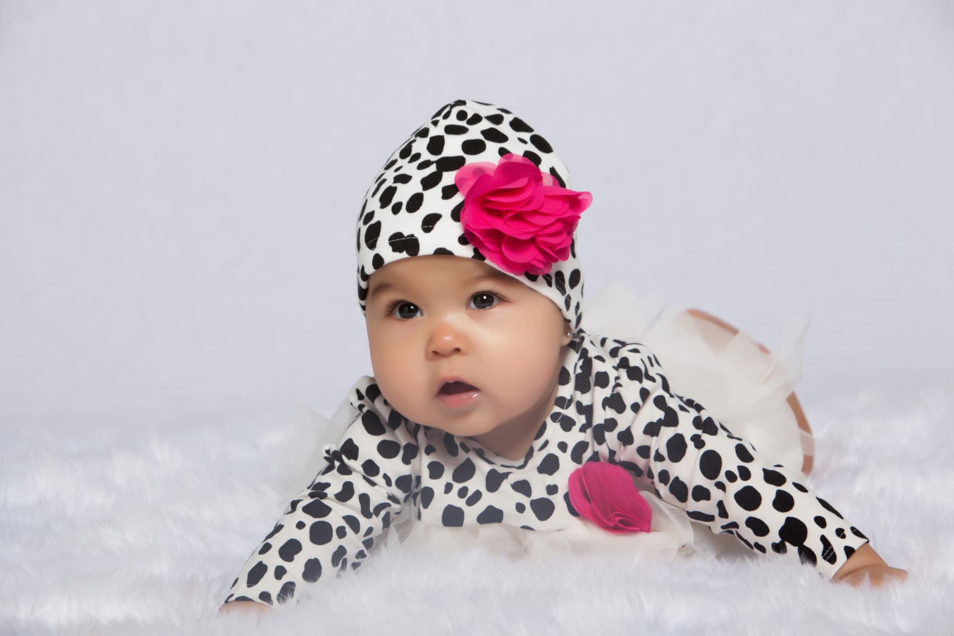 Baby girl in a spotted outfit and bright pink flower posing on a white blanket
